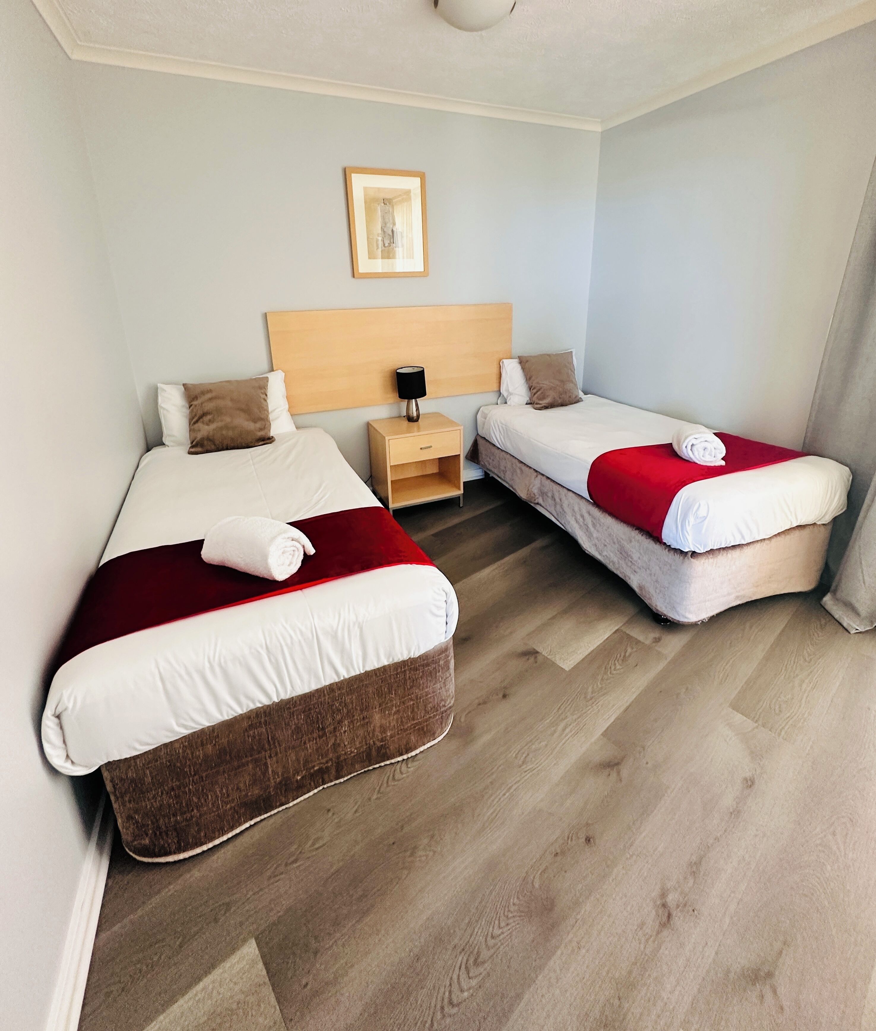 Habitación de hotel con dos camas, una mesita de noche, un cuadro enmarcado en la pared y suelo de madera.