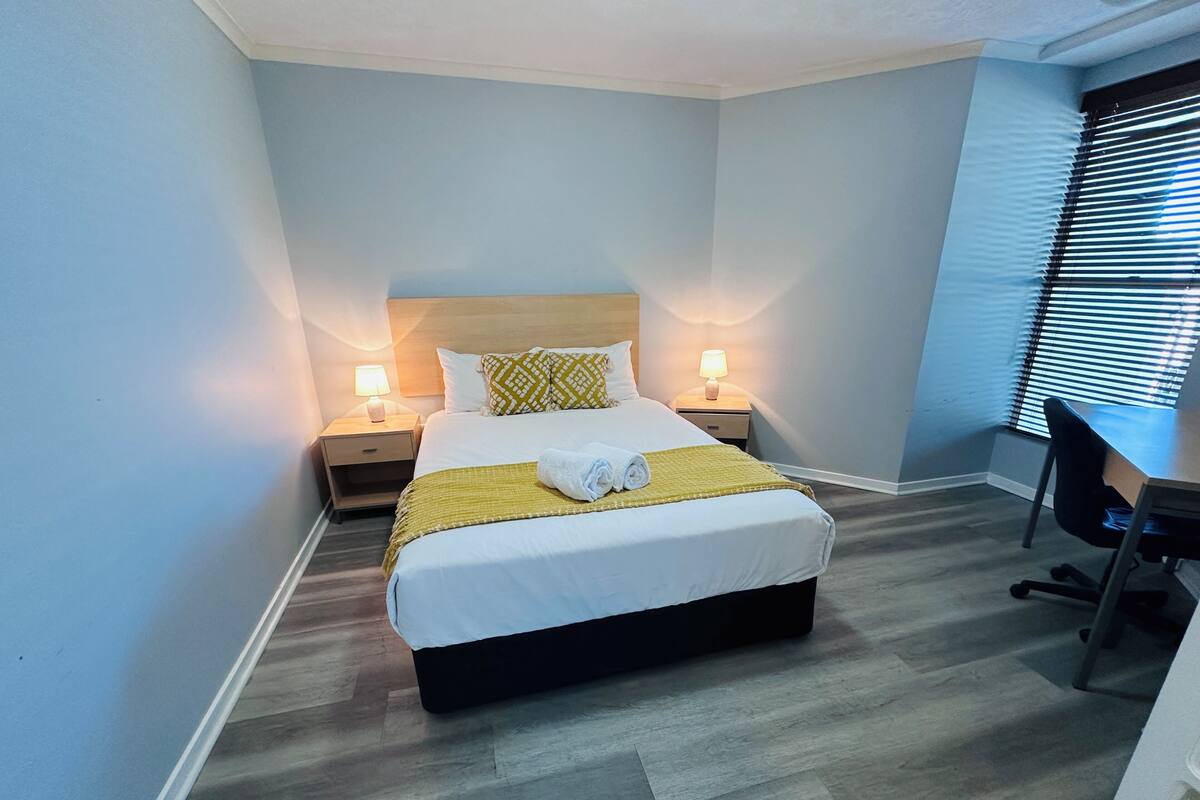 Una habitación de hotel con cama, lámparas de noche, un escritorio y una ventana con persianas.