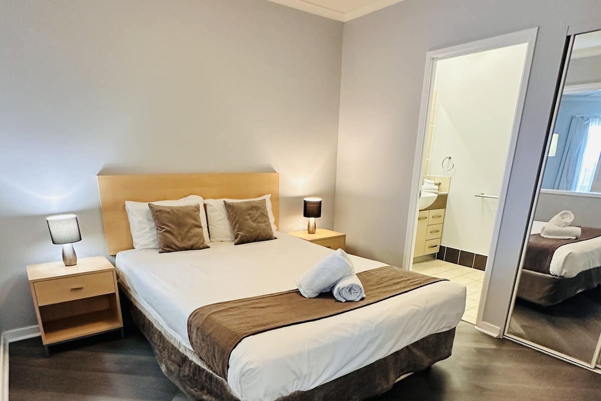 Una cama bien hecha con cabecera marrón, dos mesitas de noche con lámparas, un espejo grande y un baño visible a través de una puerta abierta.