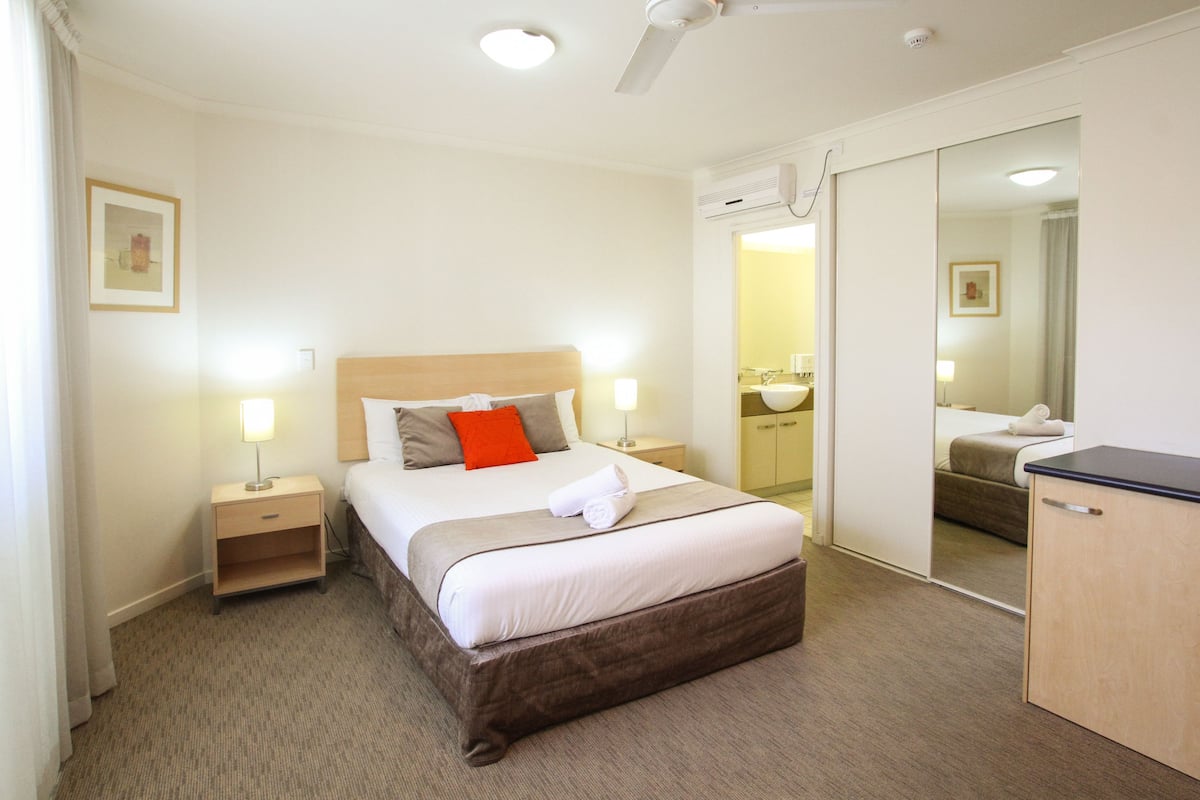 Una habitación de hotel con cama, mesitas de noche, un espejo, armario y baño visible a través de una puerta abierta.