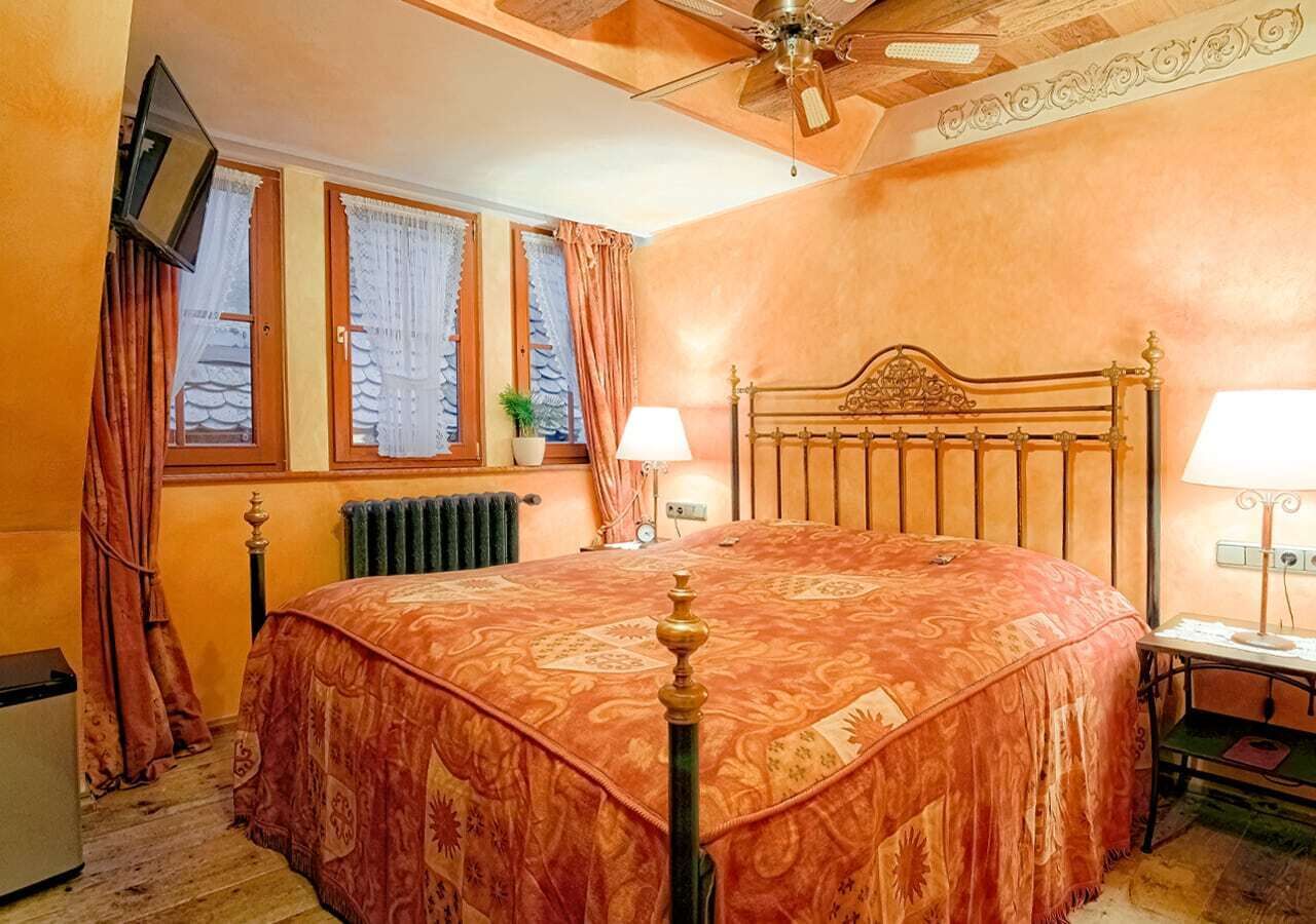 A bedroom with a large bed, two bedside lamps, a ceiling fan, a radiator, and a window with curtains.