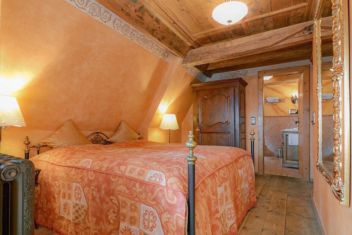 A bedroom with a large bed, two bedside lamps, a wooden ceiling, and a door leading to a bathroom.