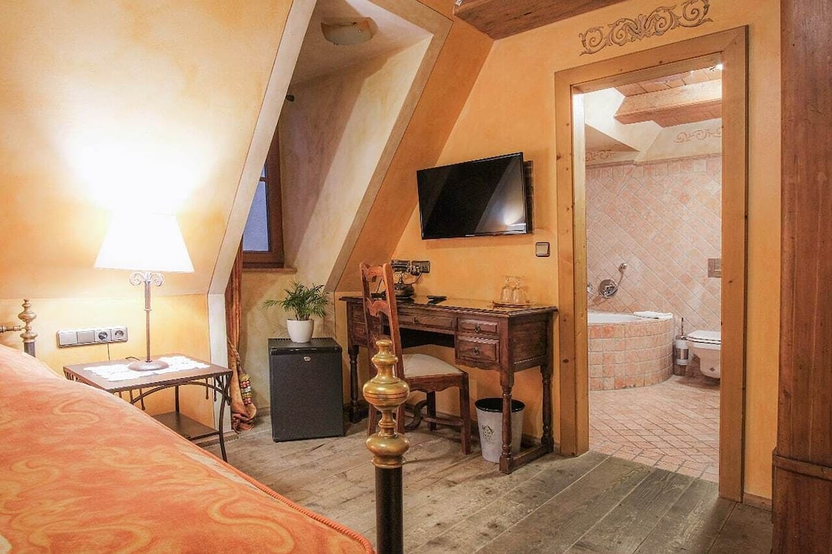 A bedroom with a sloped ceiling, a desk, a chair, a television, and a bathroom visible through an open door.