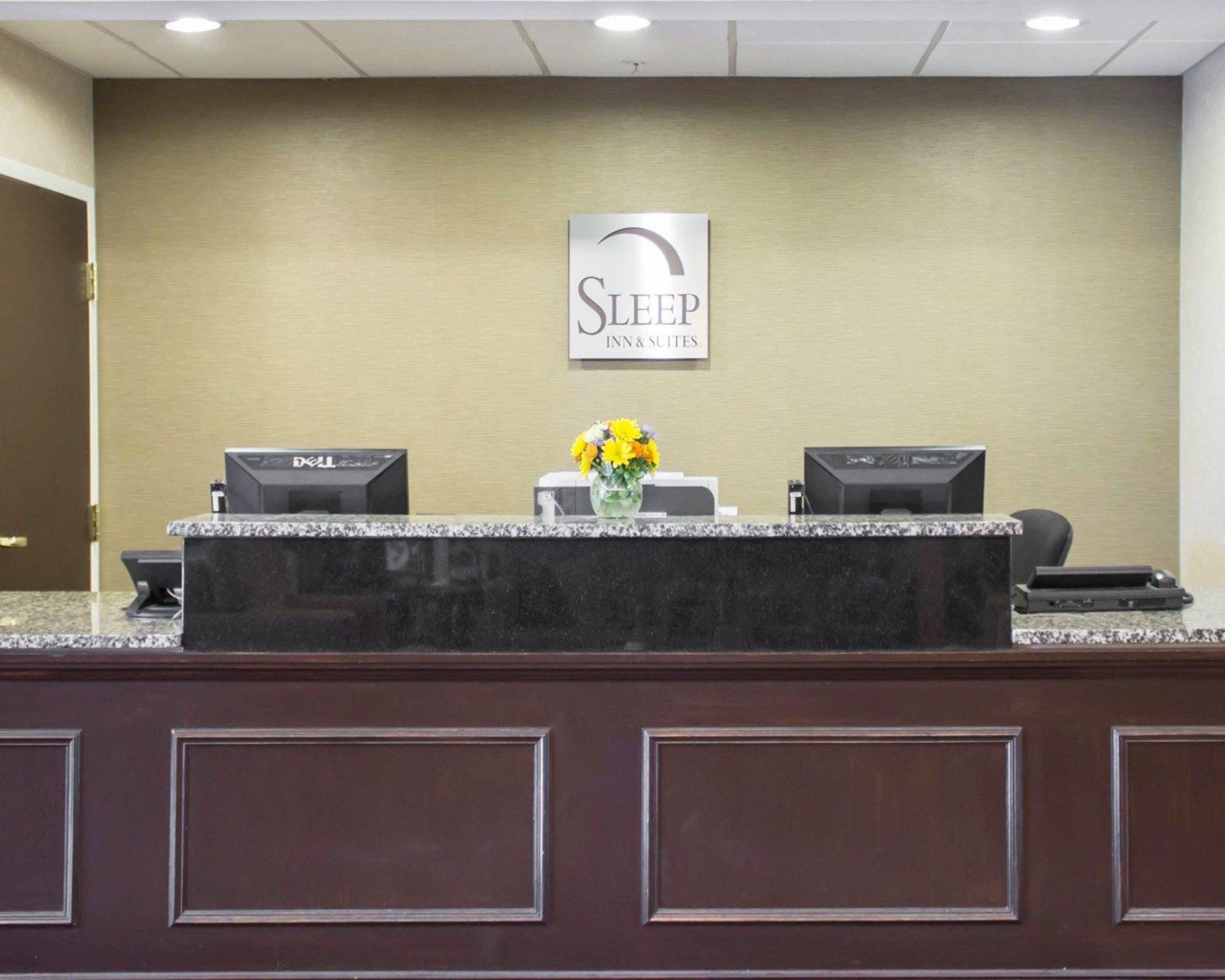 Un comptoir d’accueil sur lequel est affiché un panneau indiquant « SLEEP INN & SUITES ». On y trouve deux écrans d’ordinateur et, au centre, un vase rempli de fleurs.