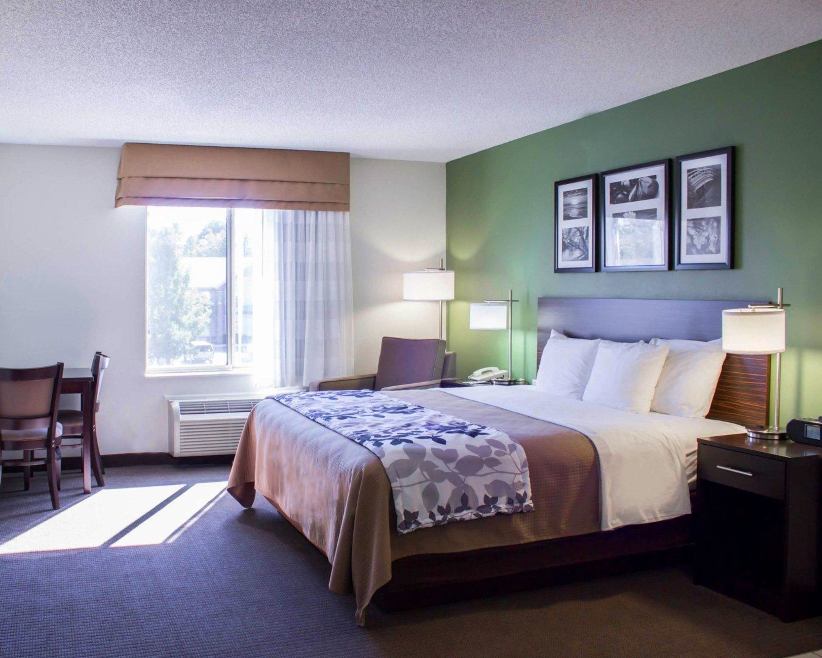 A hotel room with a bed, bedside table, lamp, chair, and a window with curtains.