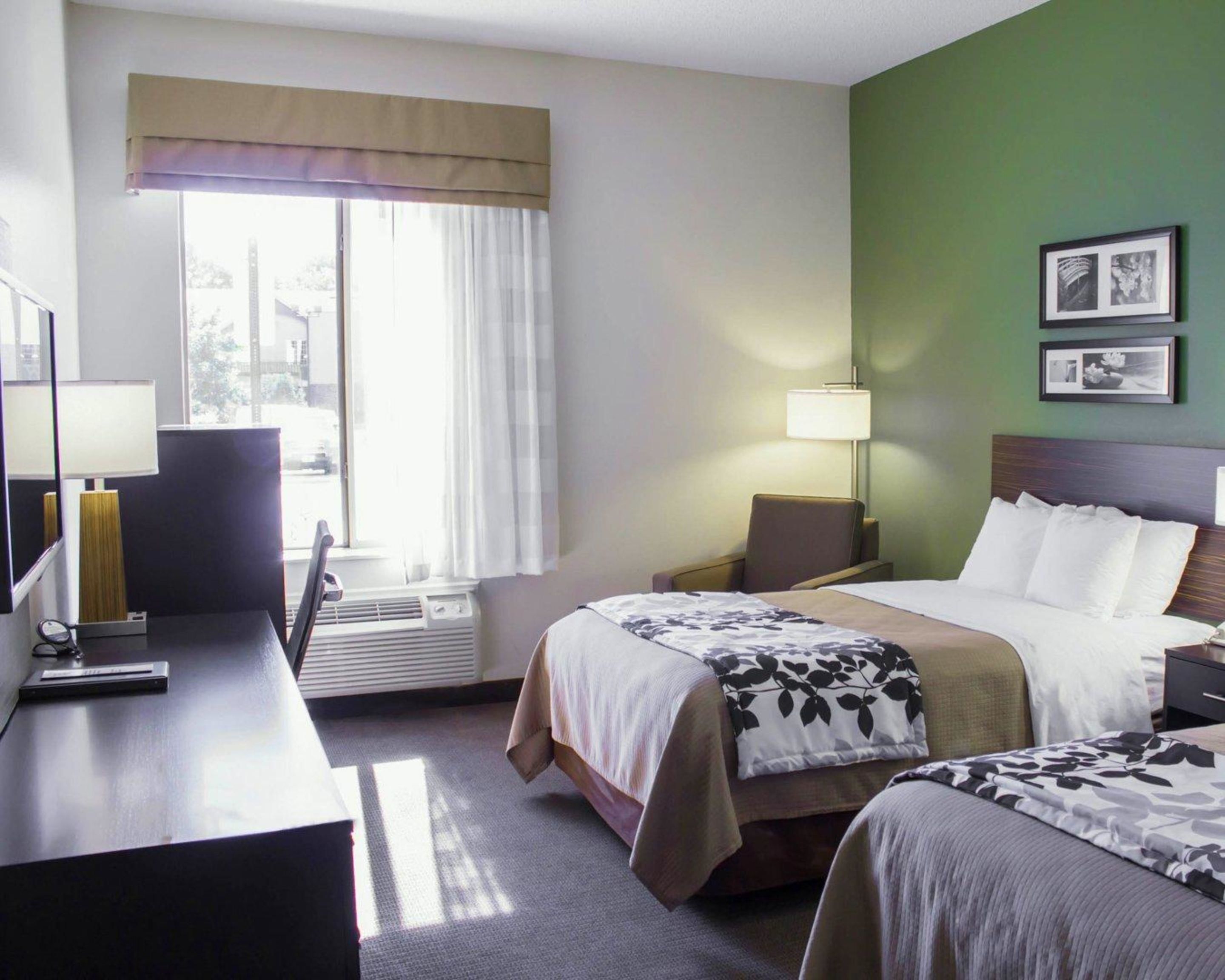 Ein Hotelzimmer mit zwei Betten, einem Schreibtisch, einem Stuhl, einem Fenster mit Vorhängen, einer Lampe und gerahmten Bildern an der Wand.