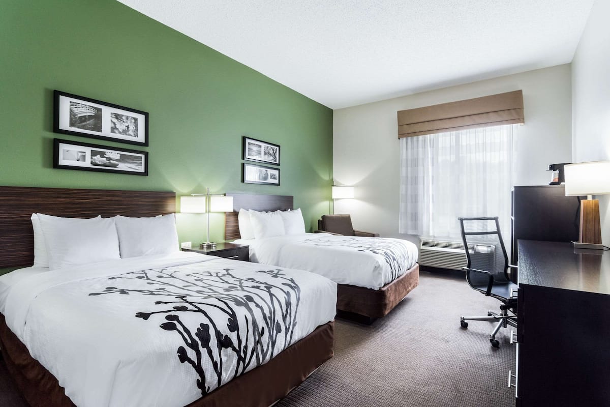 A hotel room with two beds, a desk, and a chair. There are framed pictures on the wall and a window with curtains.