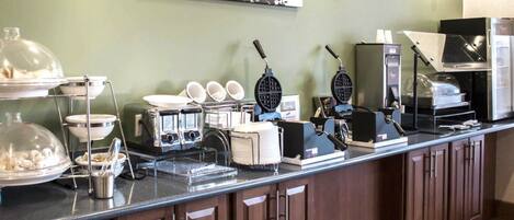 A buffet setup with various food items and appliances, including a coffee machine and a waffle maker, against a green wall with a decorative piece above.
