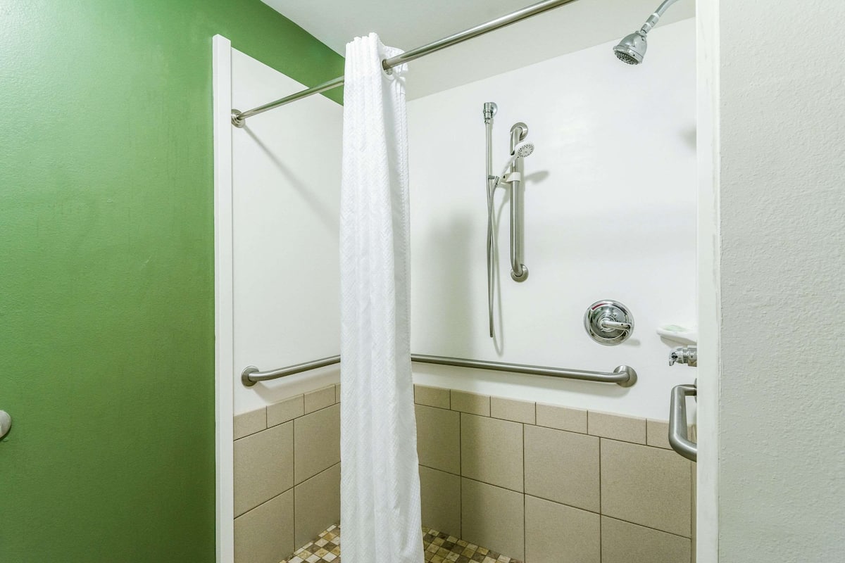 A shower area with a white curtain, grab bars, a handheld showerhead, and a wall-mounted faucet.