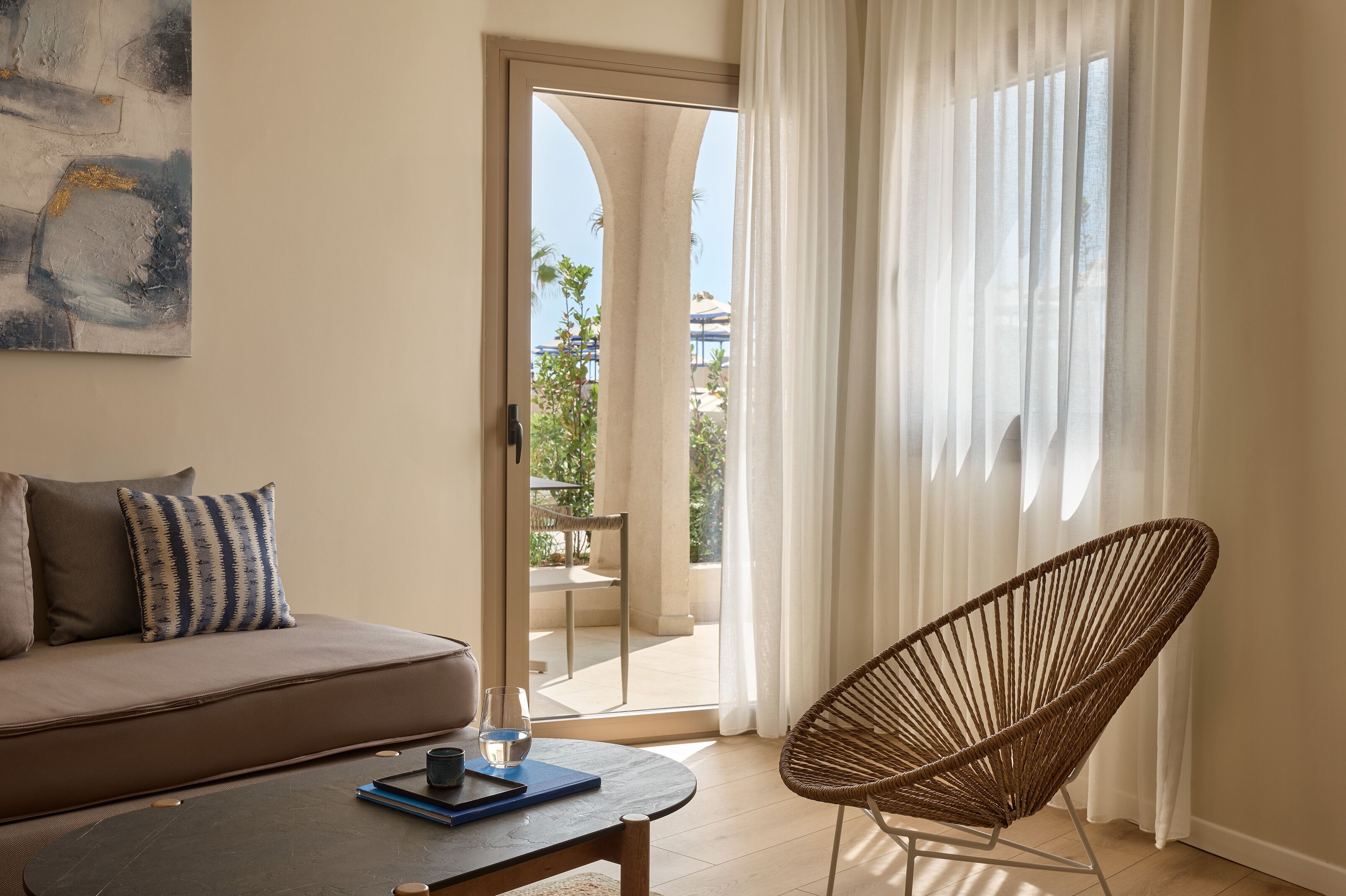A room with a sofa, a wicker chair, a table with a book and glass, and a view through a door to an outdoor area.