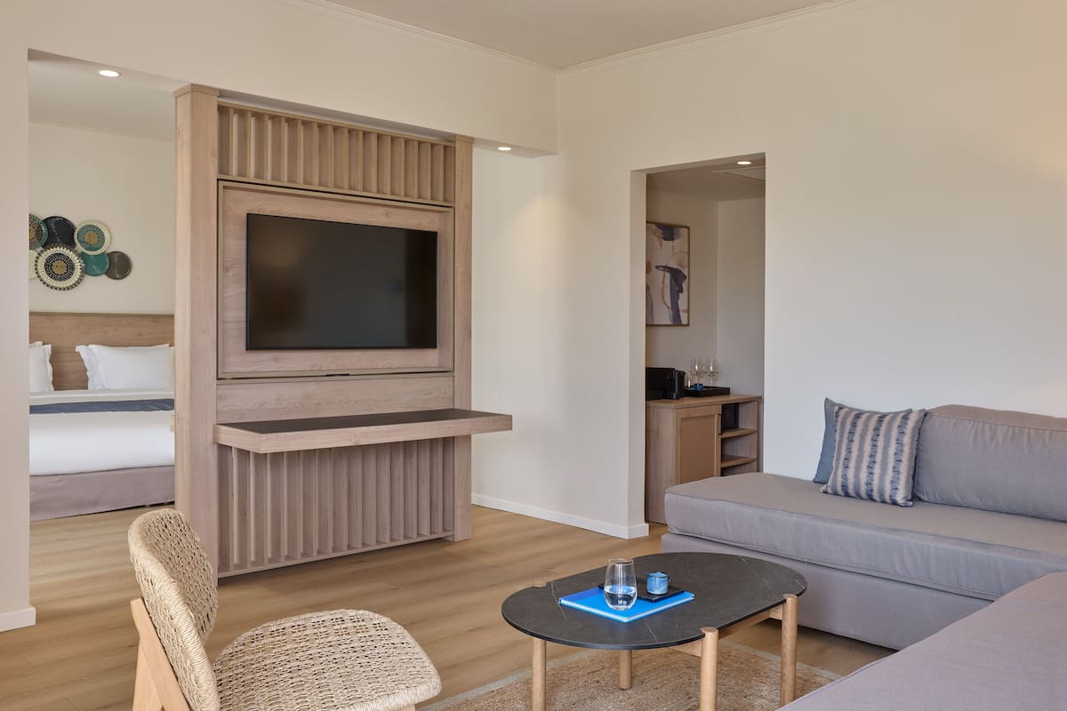 Un salon moderne avec un téléviseur à écran plat fixé sur un panneau en bois, un canapé gris, un fauteuil en osier et une petite table basse.