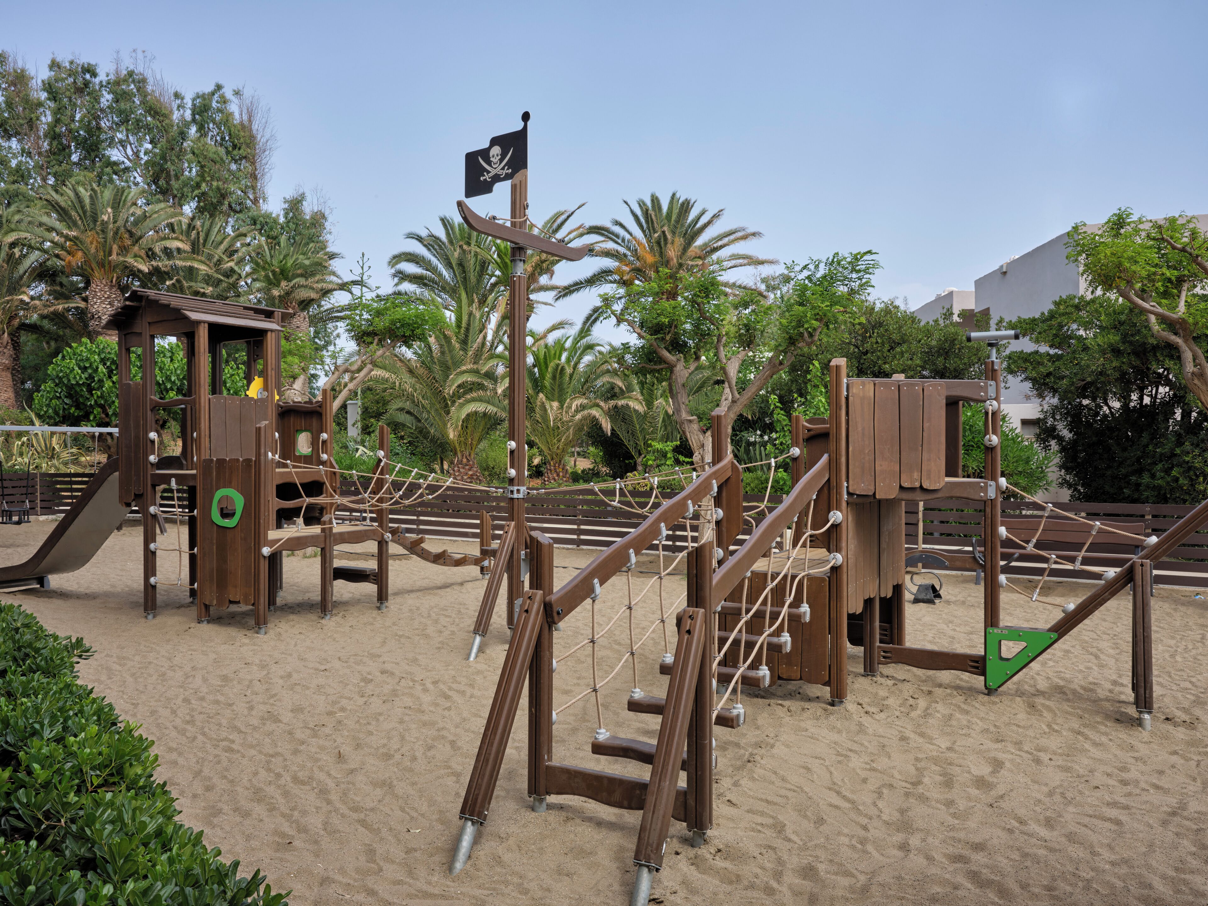 Een speeltuin met een piratenthema, inclusief een glijbaan en klimmogelijkheden, omringd door zand en groen.