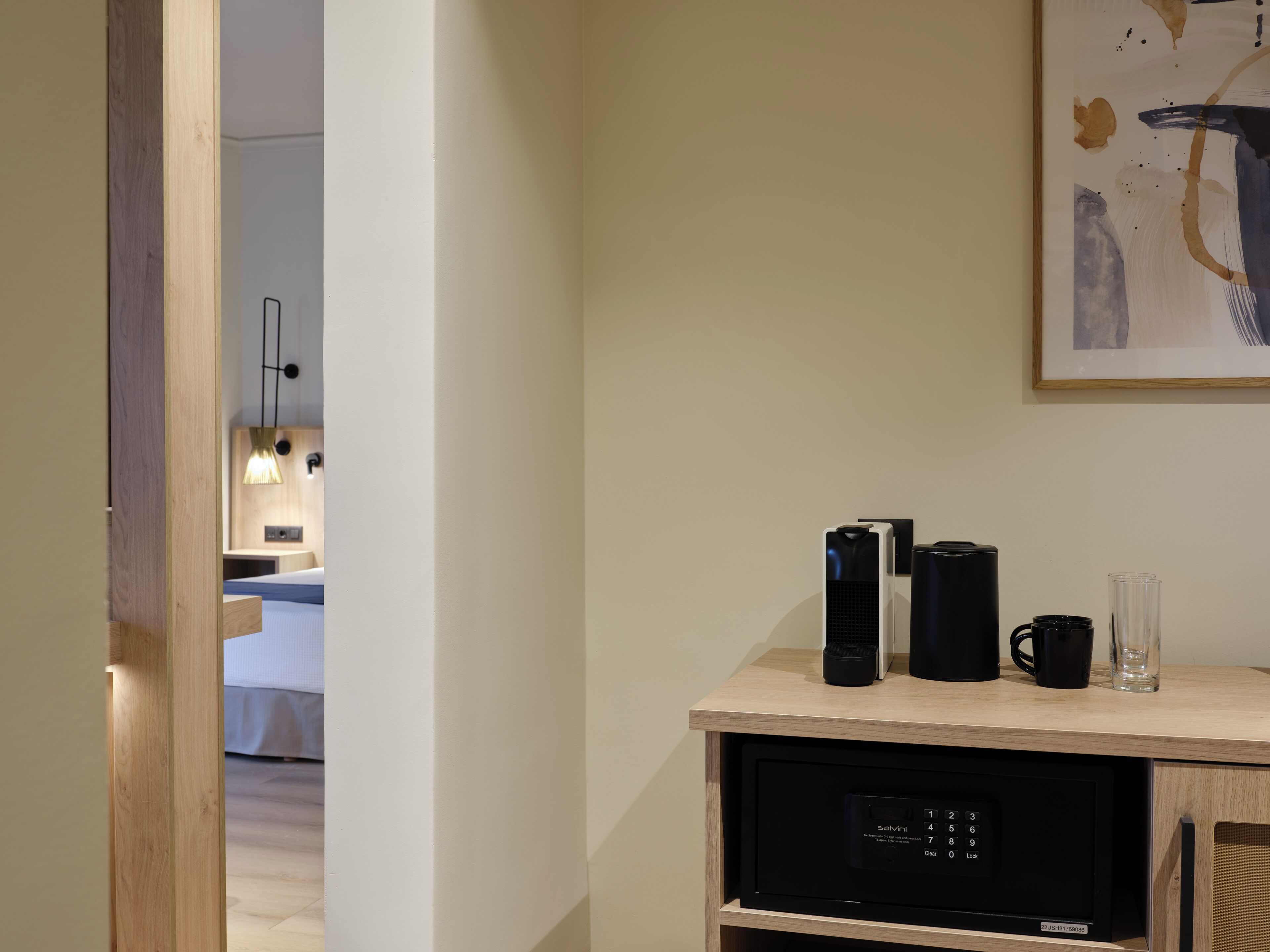 Habitación de hotel con cama, mesita de noche con cafetera, vaso y microondas.