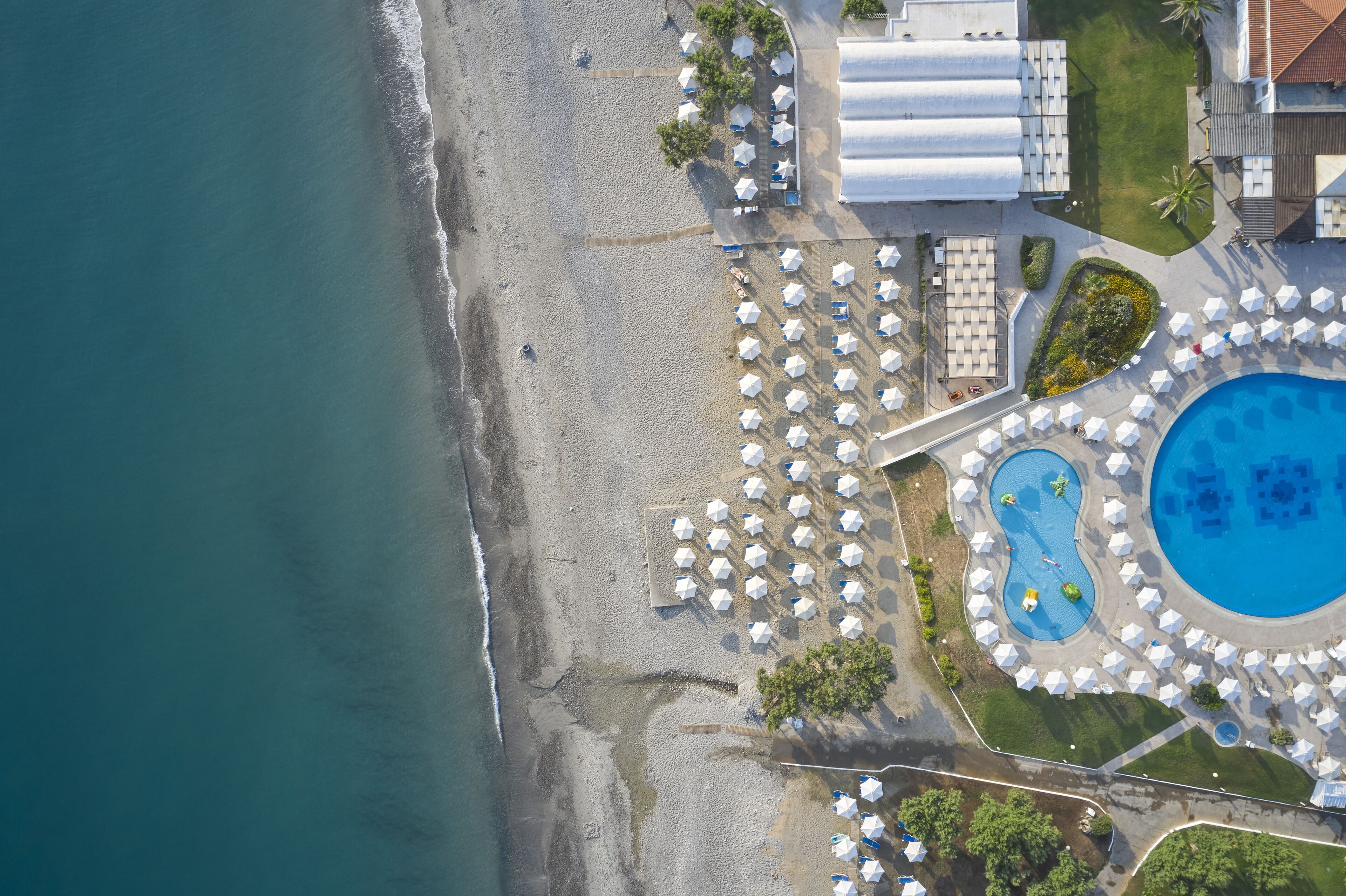 An aerial view of a beach resort with a swimming pool, beach umbrellas, and lounge chairs.