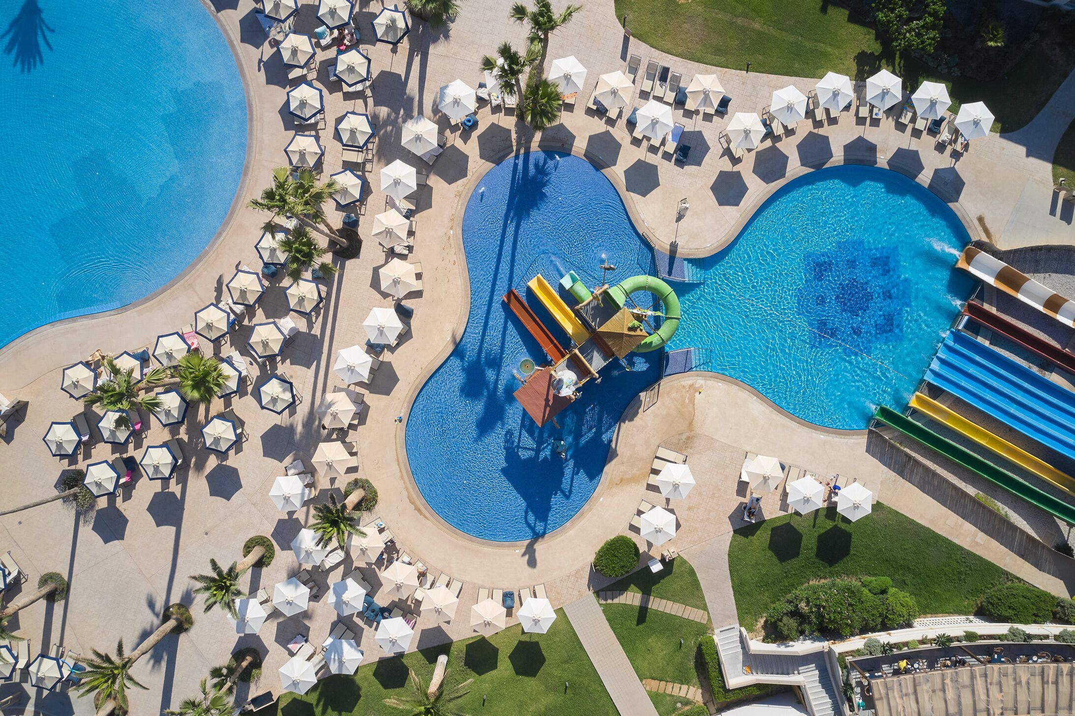 Vista aérea de un complejo turístico con una piscina grande, varias zonas de descanso con sombrillas y un tobogán de agua.