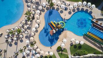 Vista aérea de un complejo turístico con una piscina grande, varias zonas de descanso con sombrillas y un tobogán de agua.