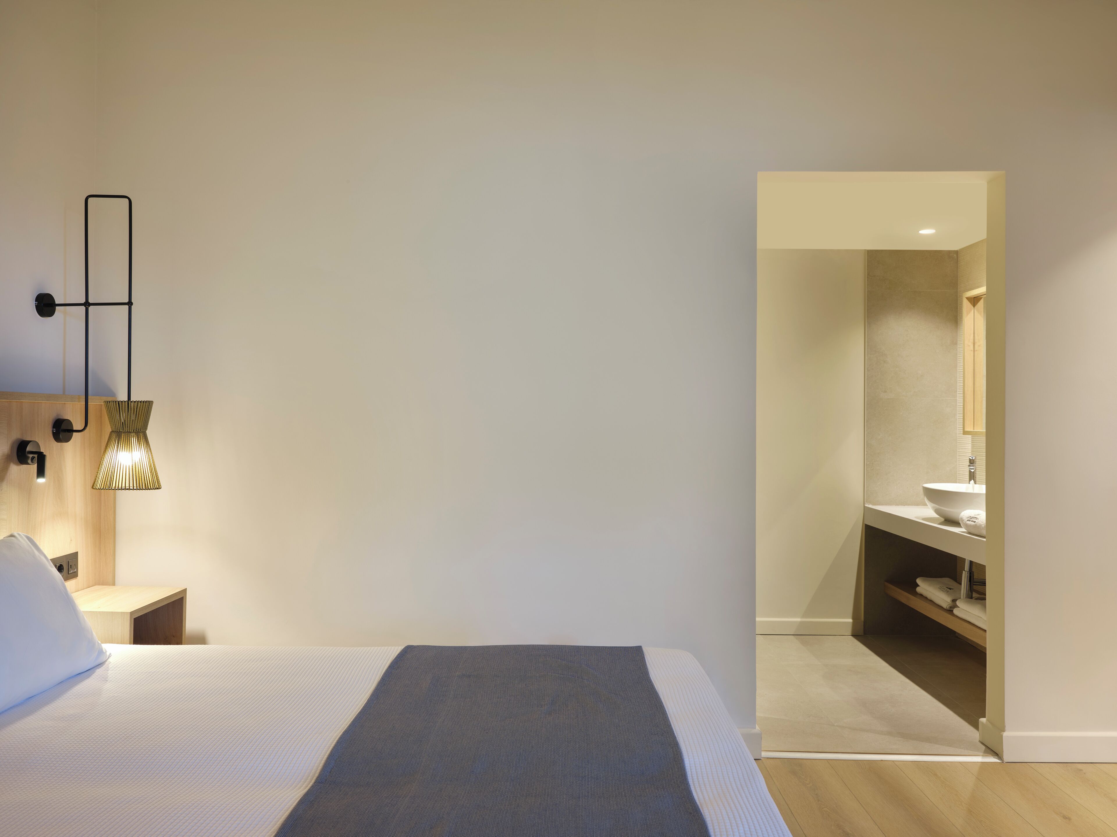 A bedroom with a bed, bedside tables, a hanging lamp, and a view into a bathroom with a sink and mirror.