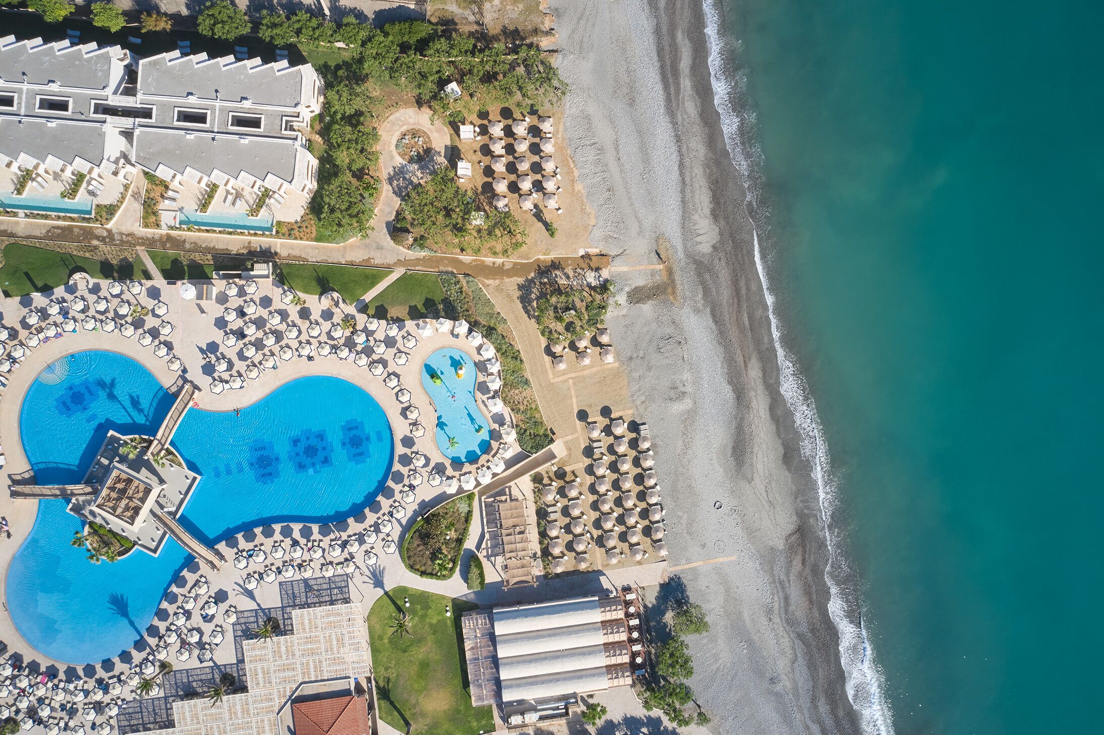 An aerial view of a resort with a large swimming pool, beach area, and multiple guest rooms.