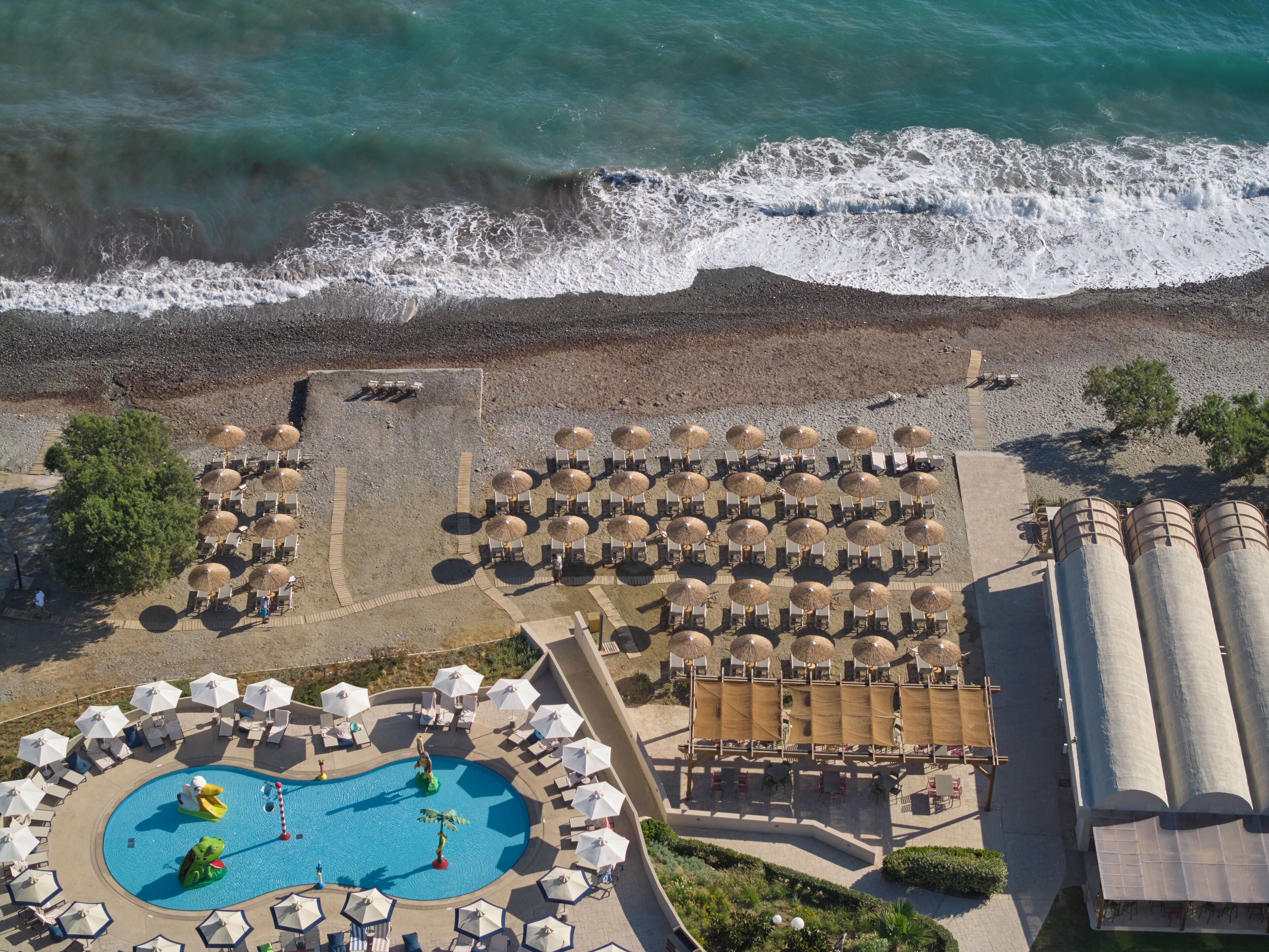 An aerial view of a resort with a swimming pool, beach umbrellas, lounge chairs, and a water slide.