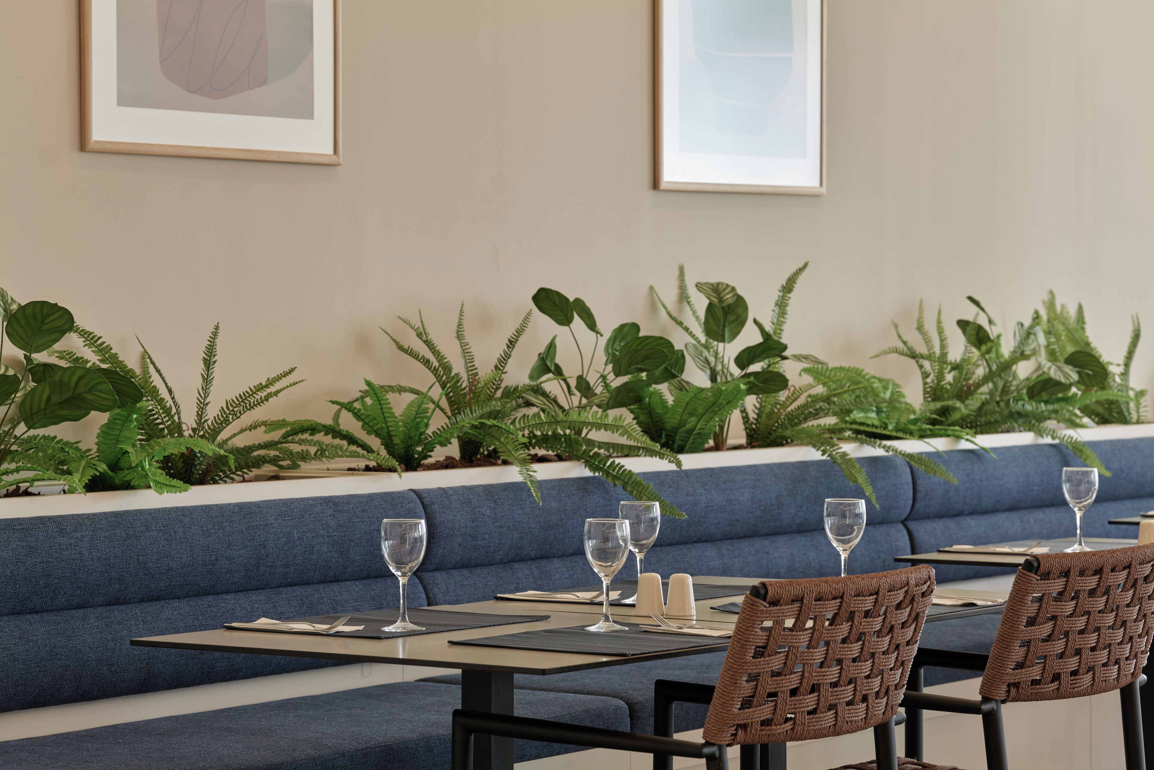 A restaurant seating area with blue cushioned benches, wicker chairs, and tables set with wine glasses and napkins.