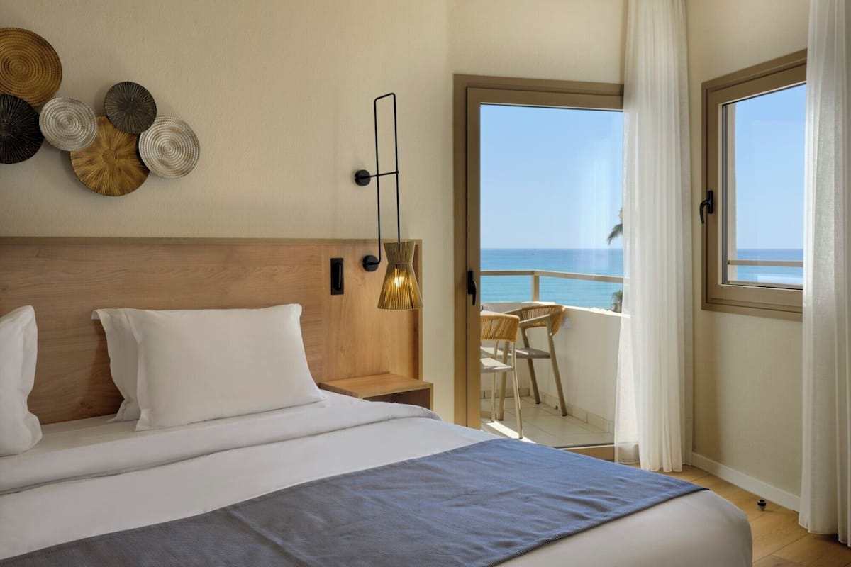 Une chambre d’hôtel avec un grand lit, une tête de lit en bois, un luminaire fixé au mur et une vue sur l’océan depuis la porte-fenêtre donnant sur le balcon.