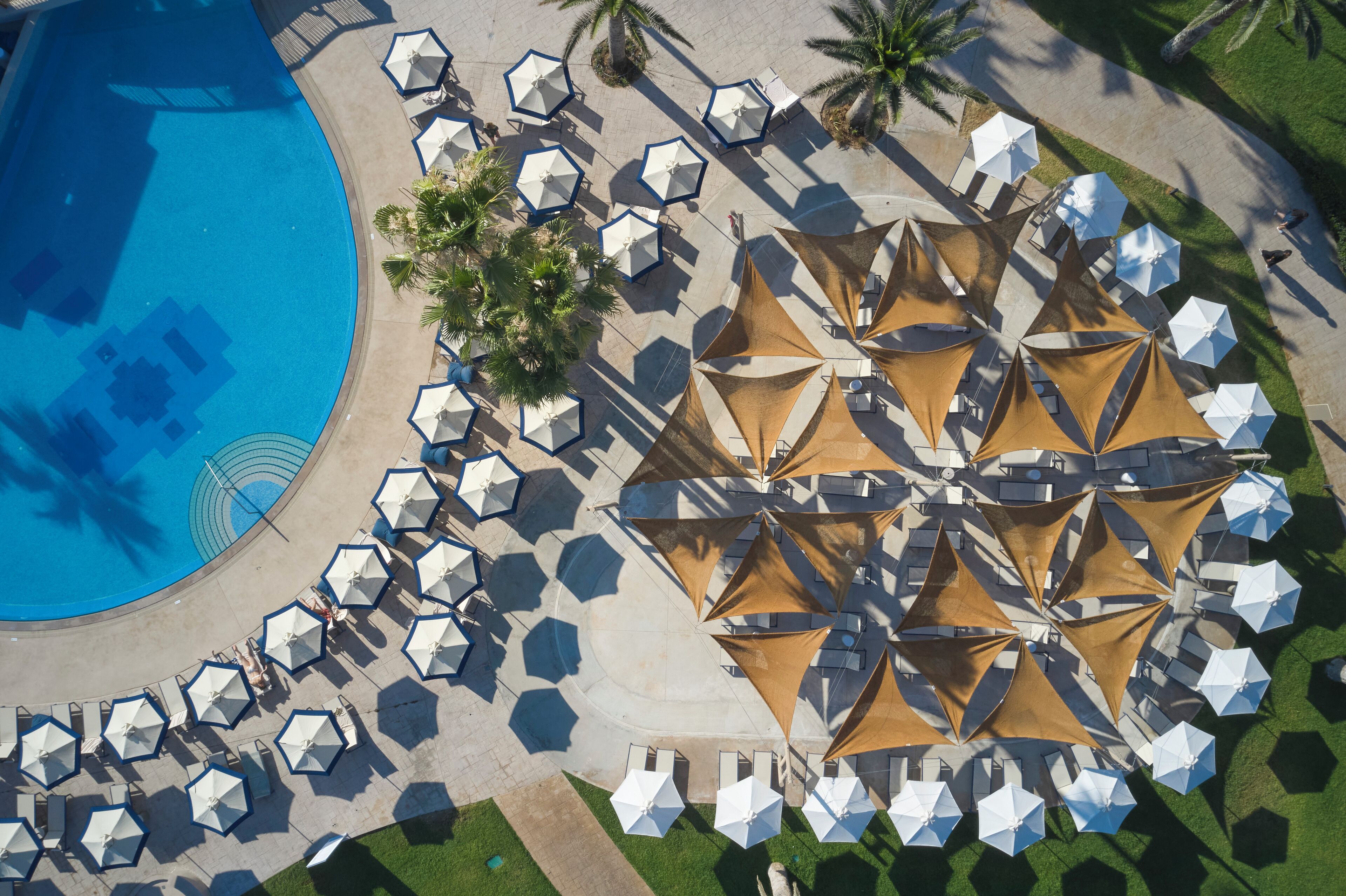 Luchtbeeld van een resort met een zwembad, zonneschermen en ligbedden die in een cirkel rond het zwembad zijn gerangschikt.
