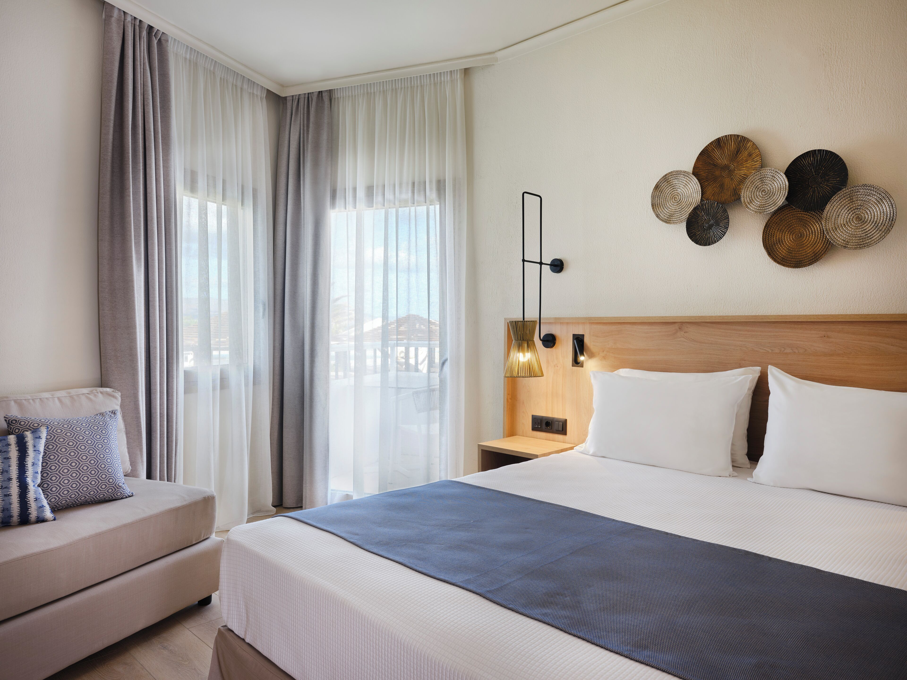 Habitación de hotel con una cama grande, una mesita de noche, un sofá y cortinas traslúcidas.