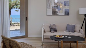 A living room with a beige sofa, a round black table with a plate of food and glasses, a woven chair, and a view of the sea through a glass door.