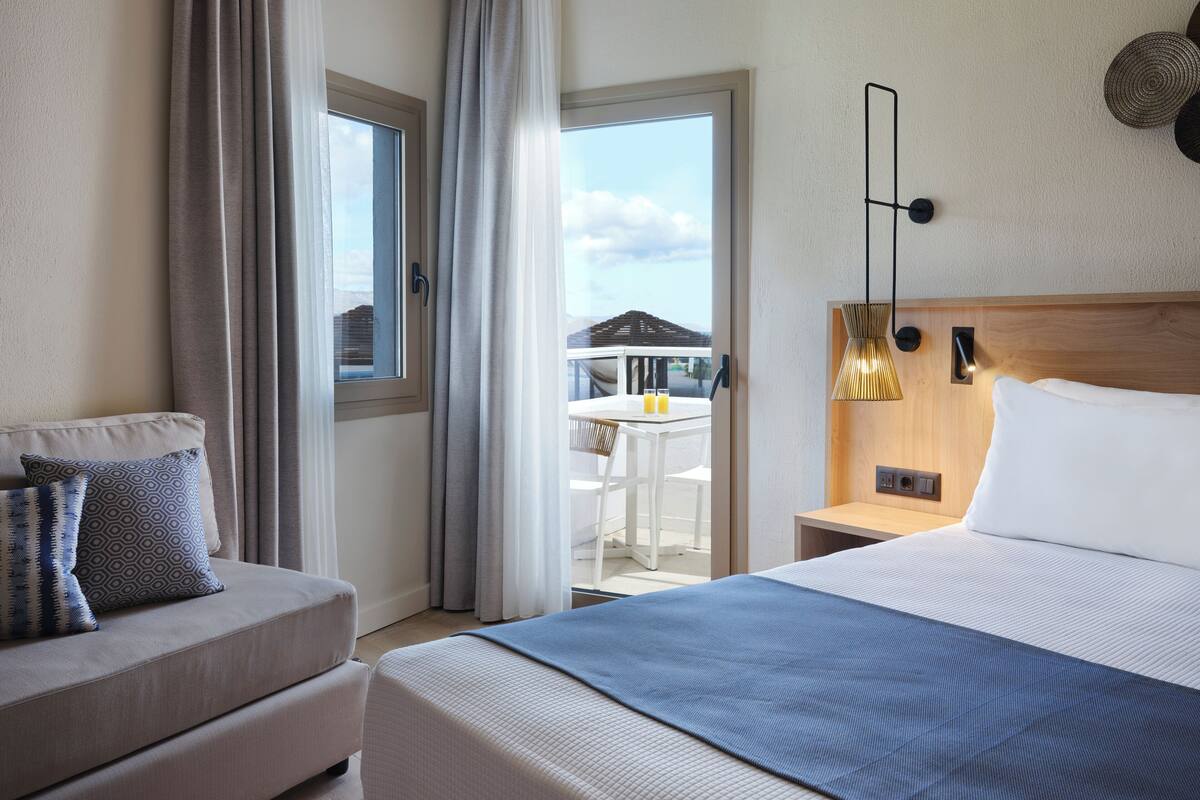 Une chambre d’hôtel comprenant un lit, un canapé, un balcon avec une table et des chaises, et donnant sur l’extérieur.