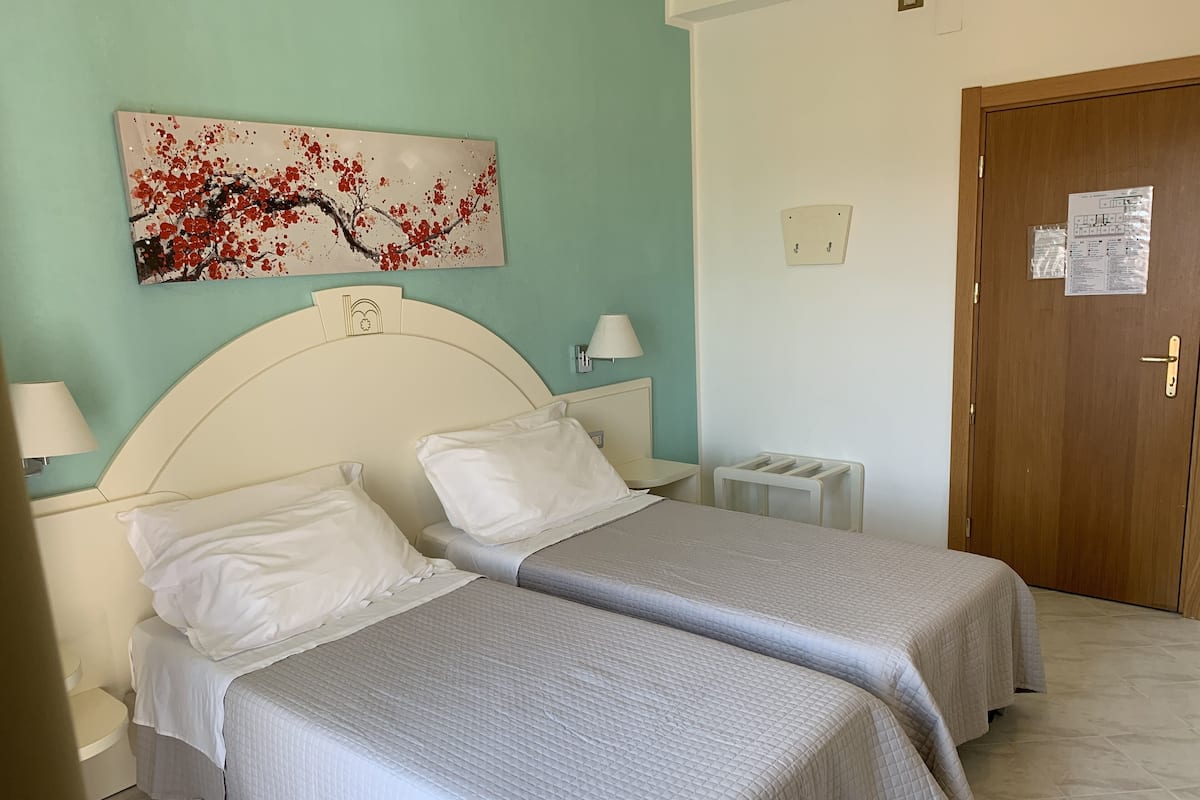 Habitación de hotel con dos camas, un cuadro en la pared, una puerta de madera y una mesita con una lámpara.