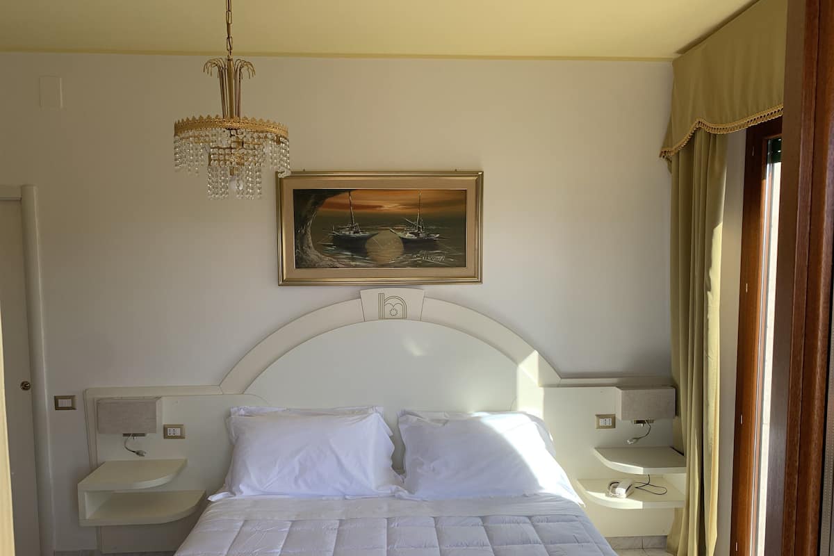 Un dormitorio con cama, mesitas de noche, candelabro, un cuadro y una ventana con cortinas.