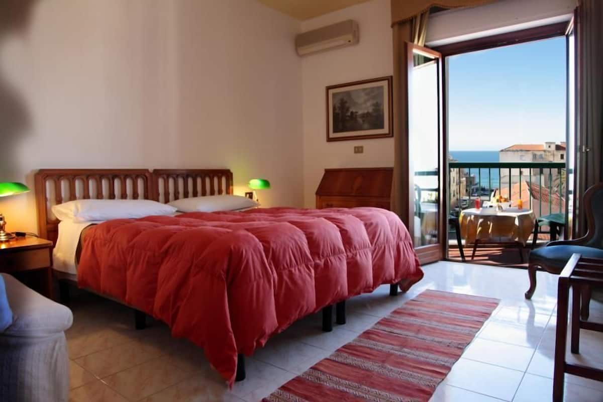 Habitación de hotel con una cama grande, un balcón con mesa y sillas, y vista al mar.