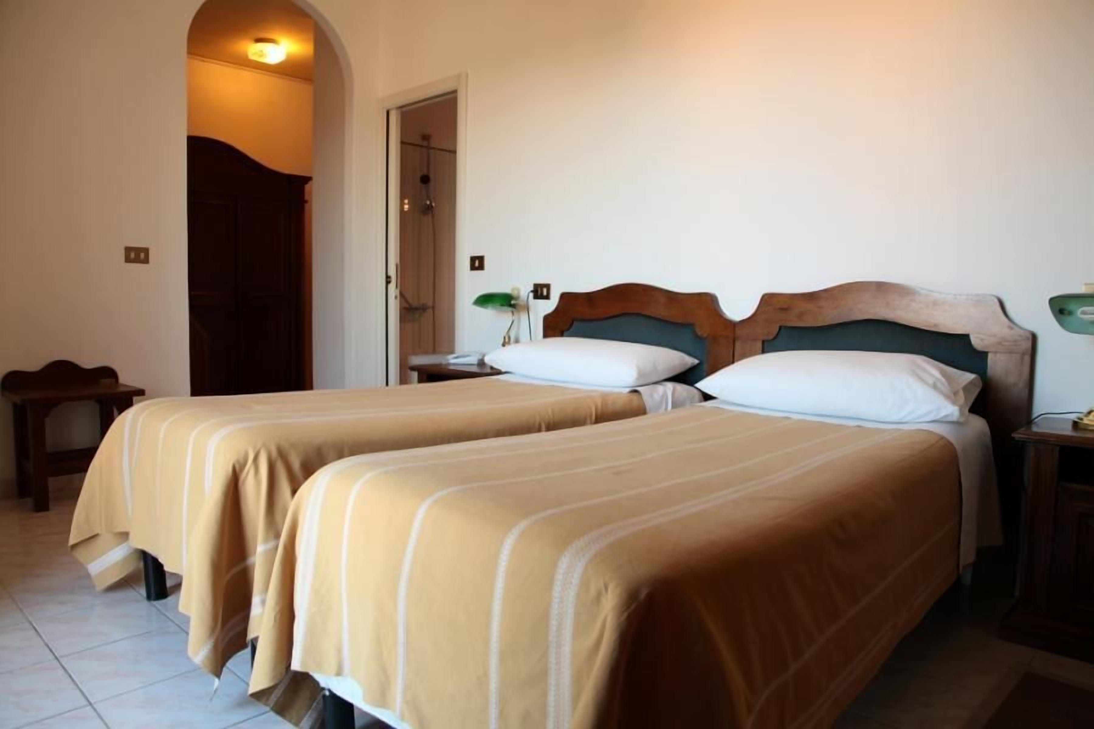 Una camera d'albergo con due letti, una testiera in legno, un comodino con una lampada e un'apertura che dà su uno spazio doccia.