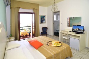 Habitación de hotel con cama, escritorio con televisor, una silla, balcón con mesa y sillas, y baño visible a través de una puerta abierta.