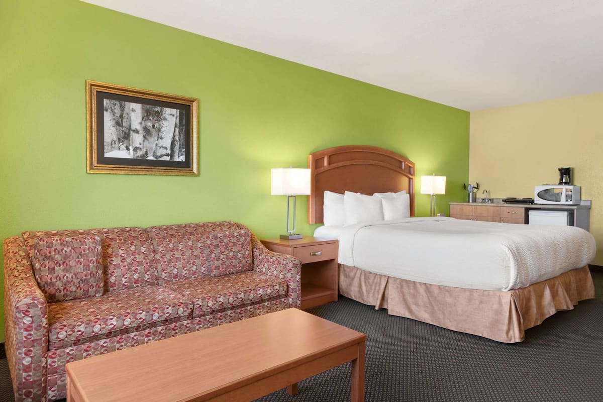 A hotel room with a bed, a sofa, a table, a nightstand, a lamp, a picture frame, and a kitchenette area.