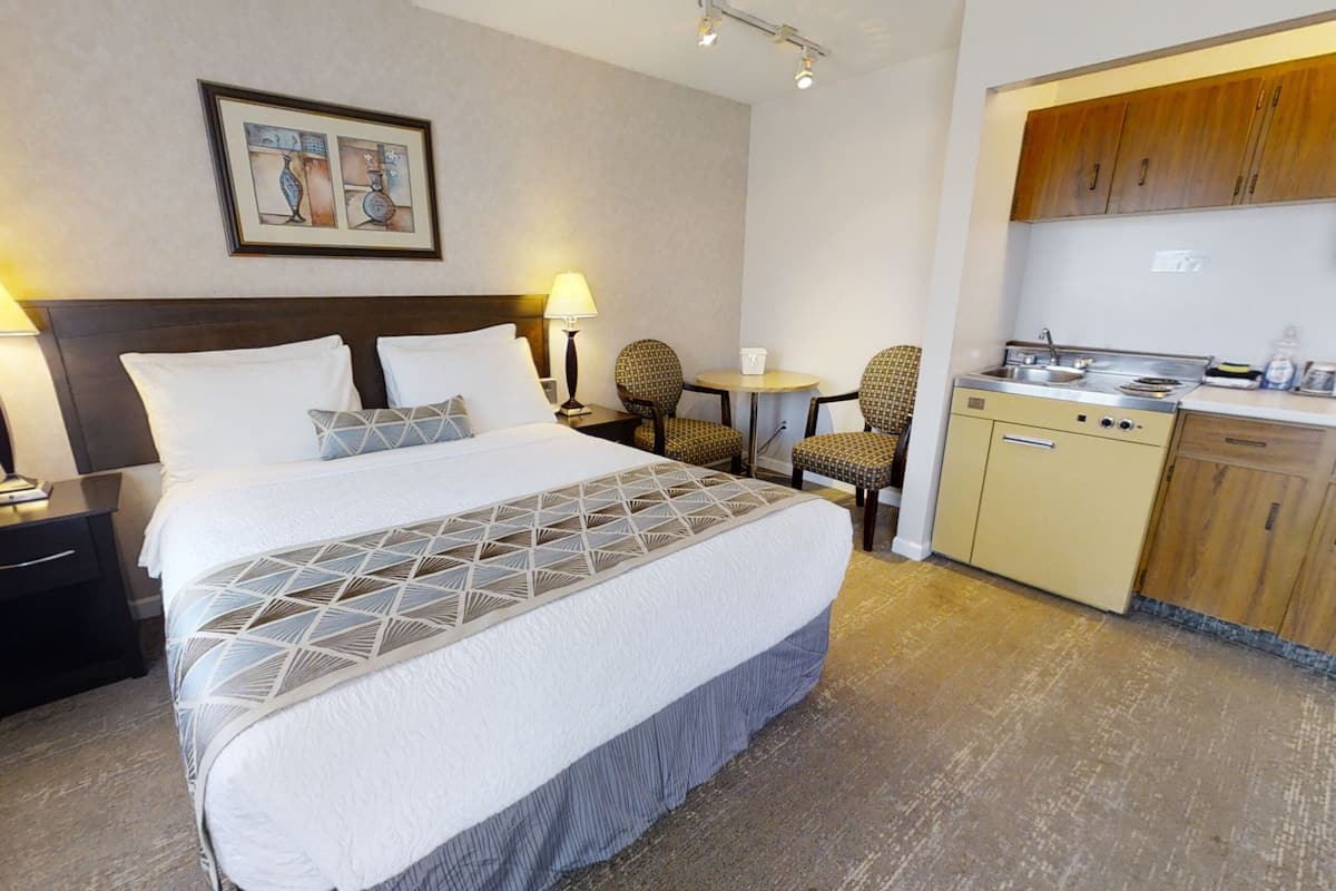 Una habitación de hotel con cama, lámparas de noche, una mesita con una silla, kitchenette y un cuadro en la pared.