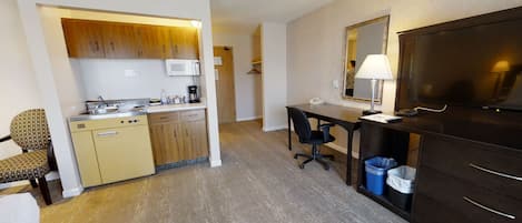 A compact hotel room with a kitchenette, a desk with a chair, a television, and a mirror.