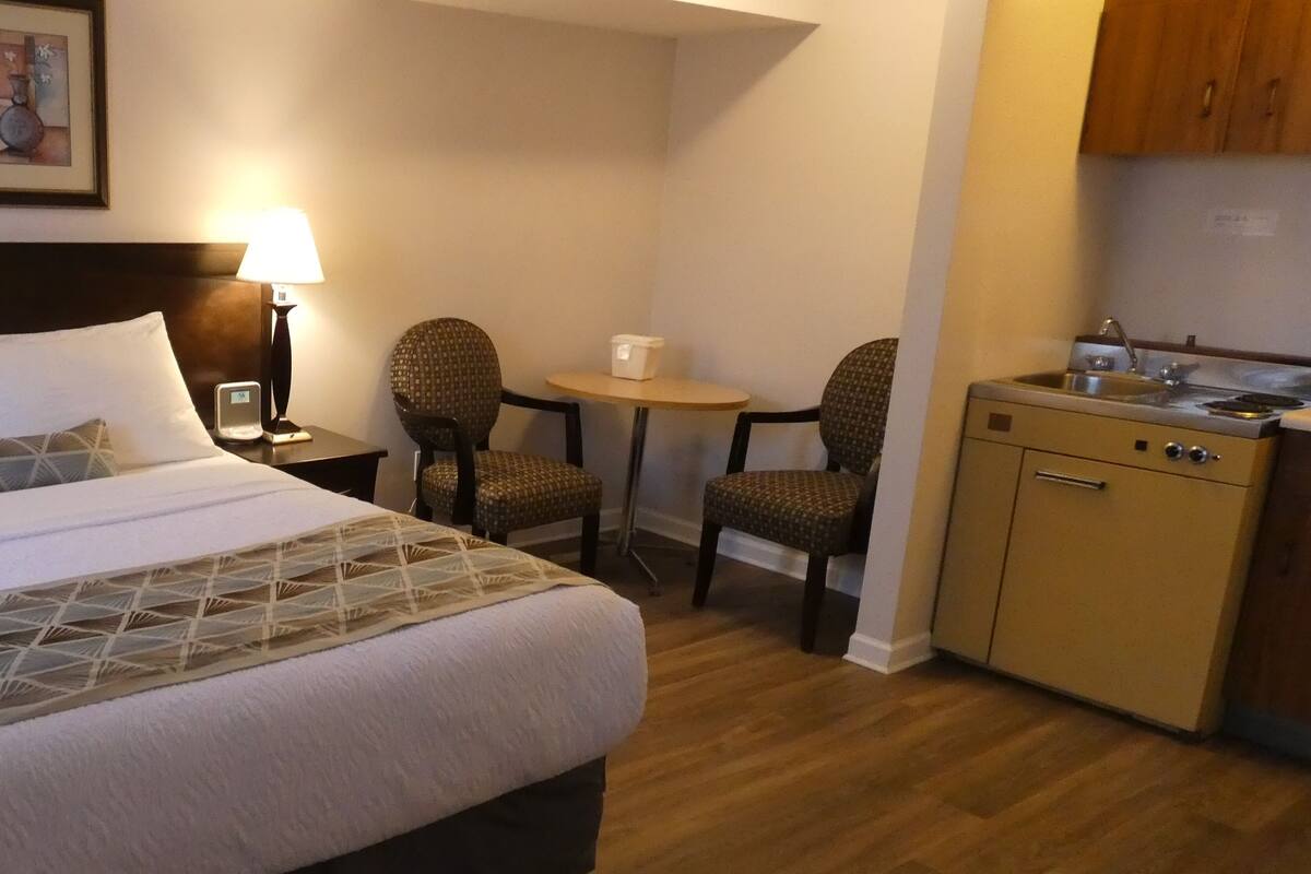 Habitación de hotel con cama, dos sillas, una mesita, escritorio y kitchenette.