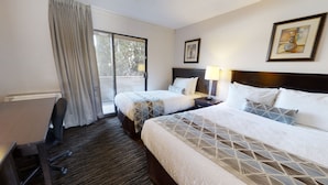 A hotel room with two beds, a desk, and a chair. There is a window with curtains, a nightstand with a lamp, and two framed pictures on the wall. - Recreation Inn & Suites (Kelowna)