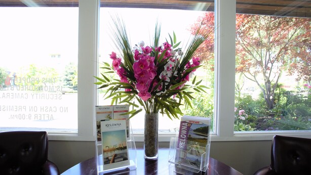 A table with a vase of flowers, brochures, and a window with a view of trees. - Recreation Inn & Suites (Kelowna)
