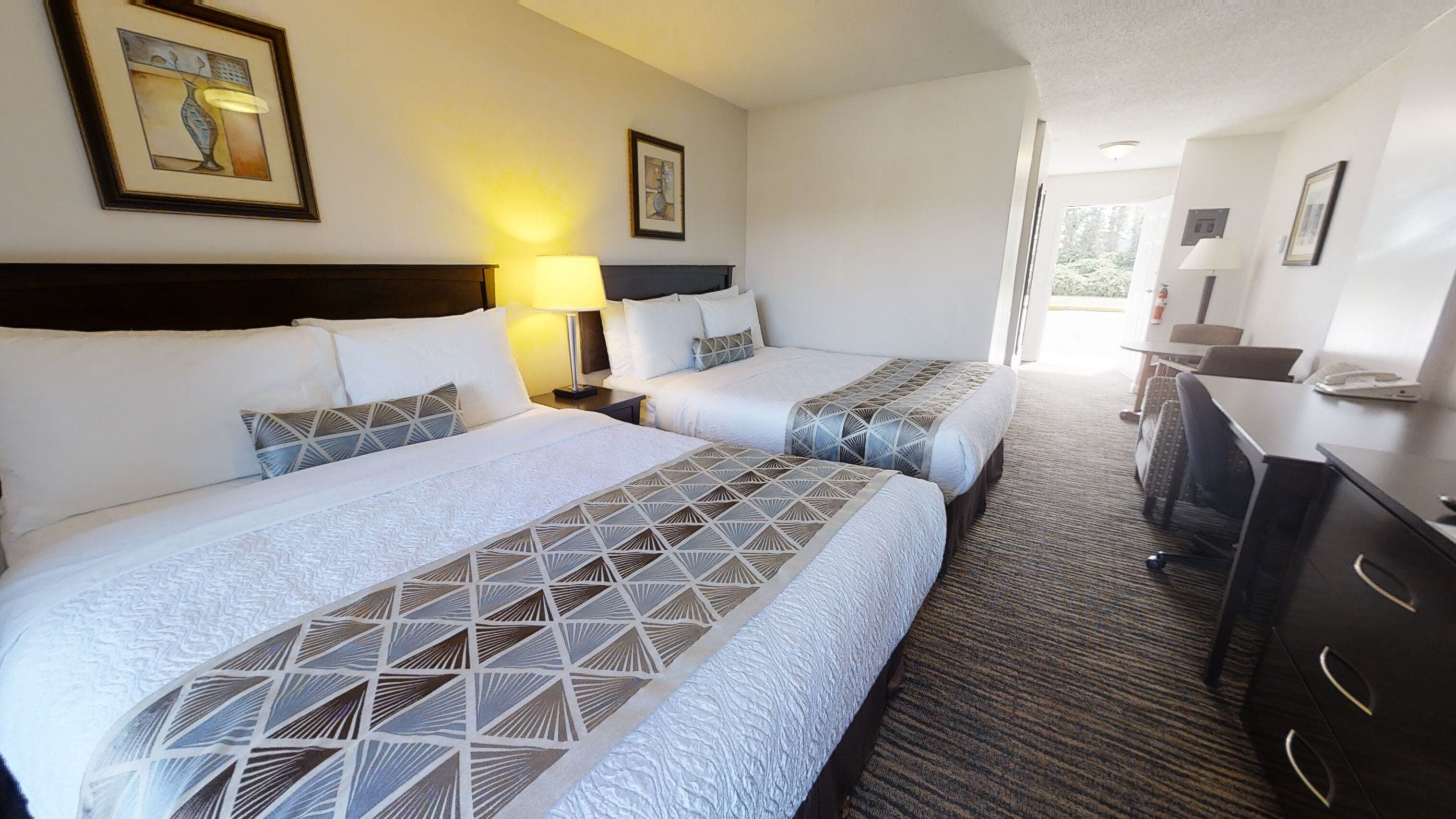 Habitación de hotel con dos camas, un escritorio y una silla. Se observan cuadros en las paredes y una lámpara en la mesita de noche.