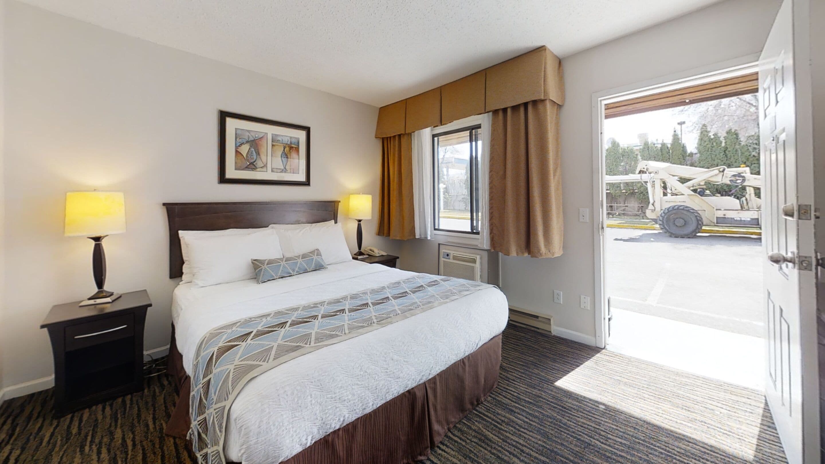 A hotel room with a large bed, two bedside lamps, a window with curtains, a framed picture on the wall, and a view of the outside through an open door.
