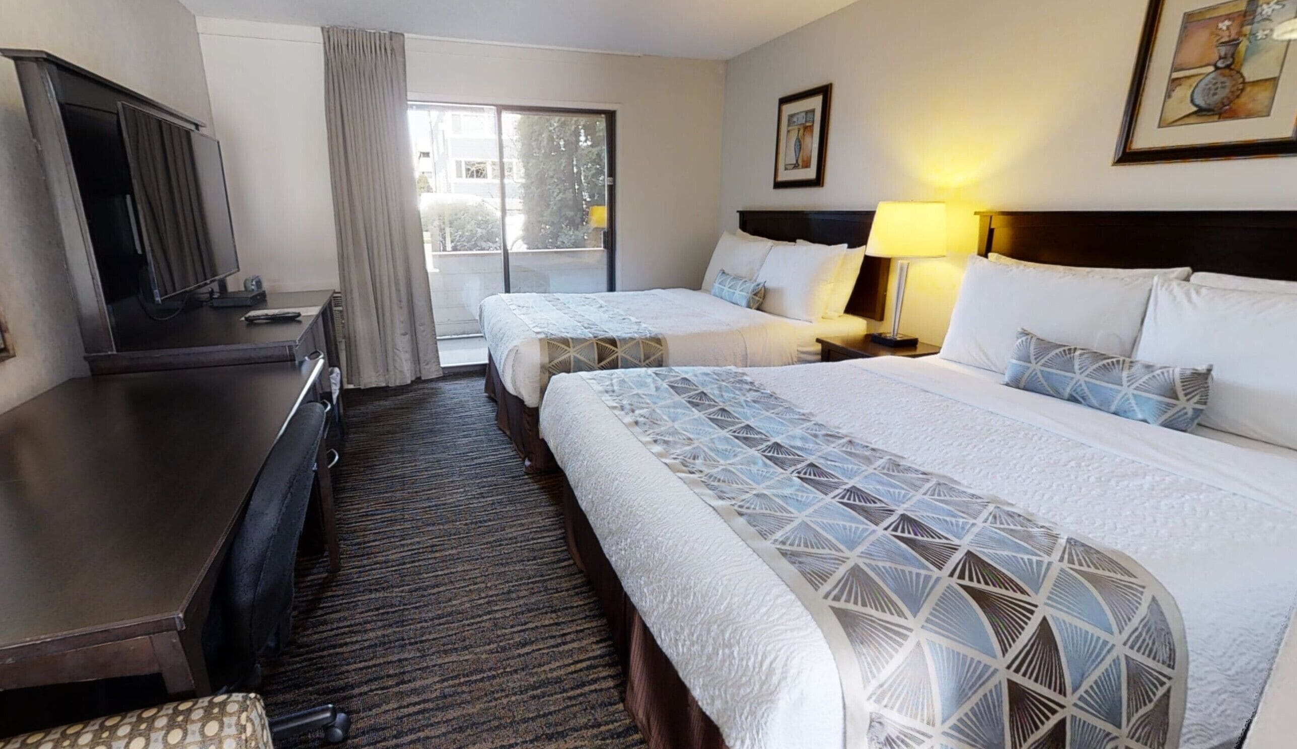 Habitación de hotel con dos camas, un escritorio, una silla, una ventana con cortinas y un cuadro en la pared.