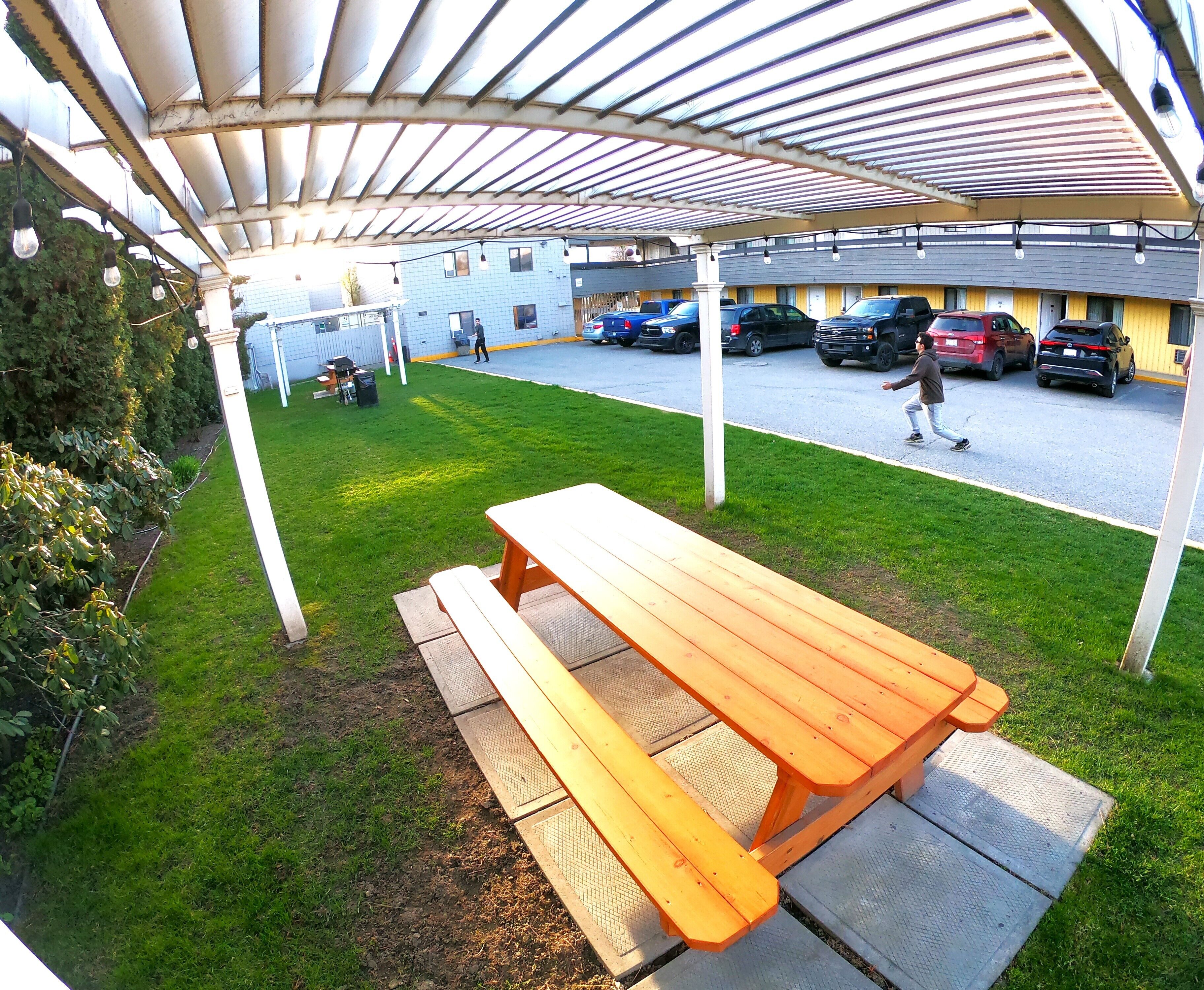 Un área de picnic techada con una mesa y bancos de madera, un estacionamiento con varios coches, y un edificio al fondo.