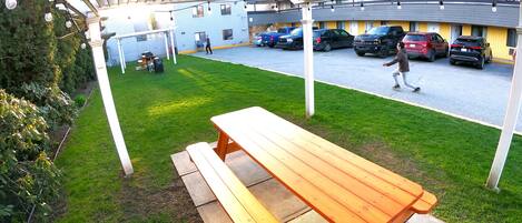 Un área de picnic techada con una mesa y bancos de madera, un estacionamiento con varios coches, y un edificio al fondo.