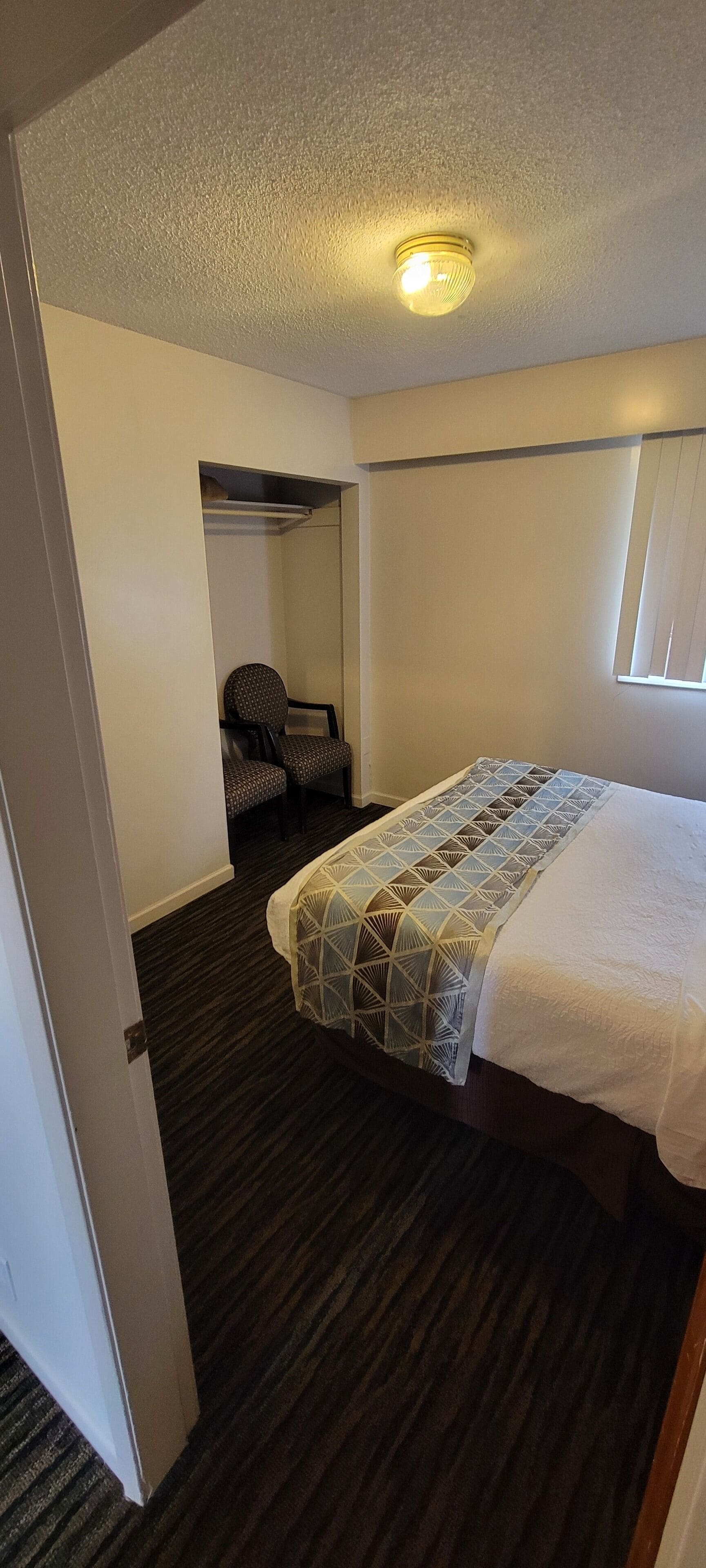 A hotel room with a bed, a chair, a closet, and a window.