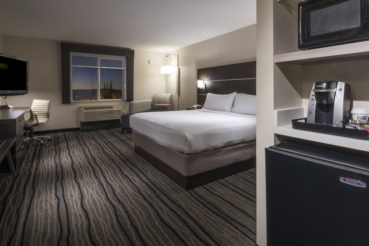 A hotel room with a bed, a desk with a chair, a television, a window with a city view, a minibar, and a coffee machine.