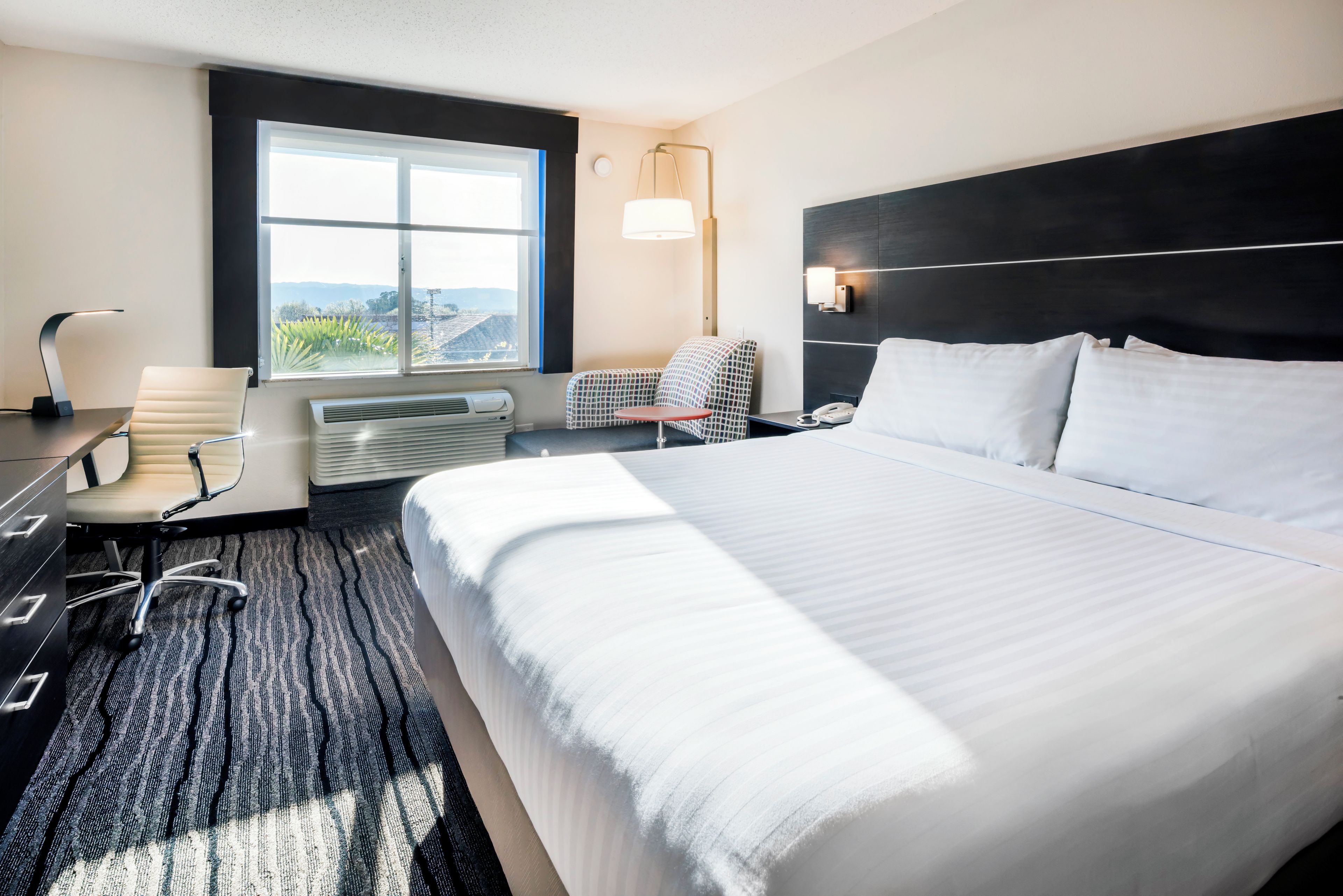 Habitación de hotel con una cama grande, un escritorio con silla, una mesita, una lámpara y una ventana con vistas.