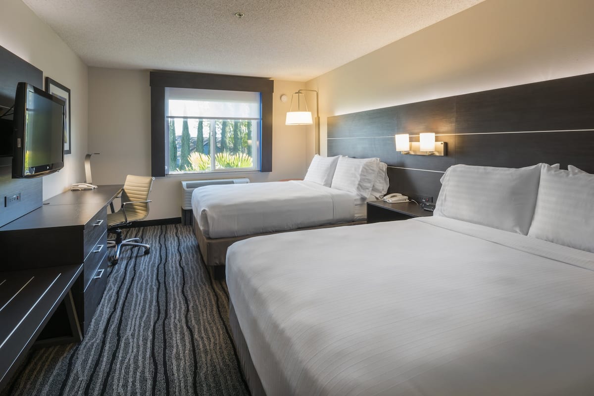 A hotel room with two beds, a desk, a chair, a TV, and a window with a view of trees.