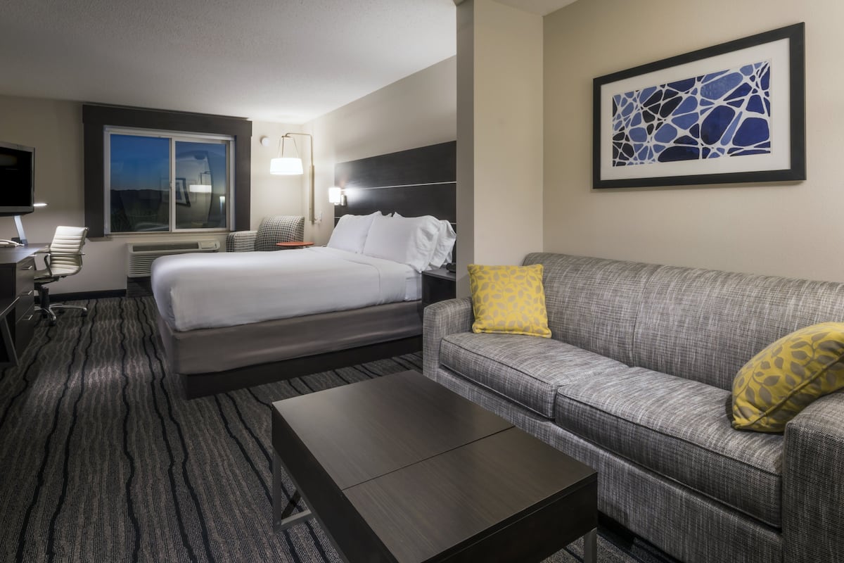 A hotel room with a bed, a gray sofa with a yellow pillow, a coffee table, a desk with a chair, a flat-screen TV, and a framed abstract art piece on the wall.